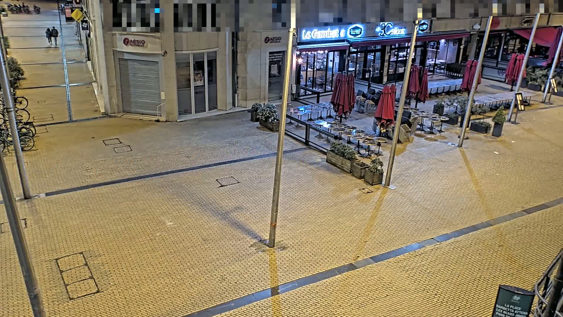 At night, an empty paved plaza with bistro tables and chairs under closed red umbrellas sits outside a closed restaurant, adjacent to a street with parked bicycles.