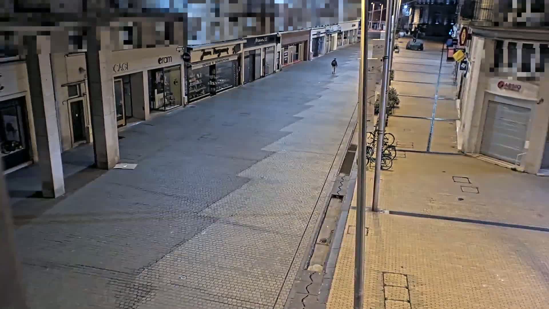 A mostly empty street at night, lined with shops, with a single pedestrian and some bicycles visible.