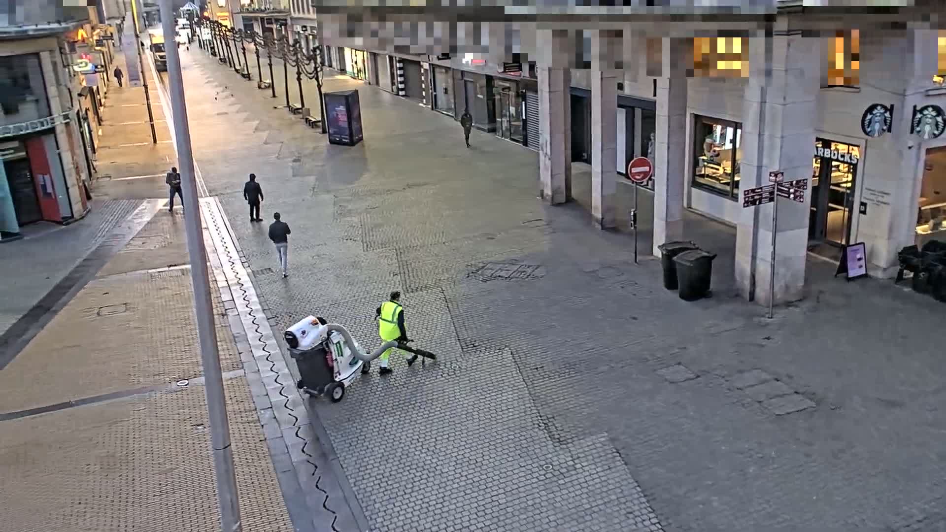 A street cleaner uses a leaf blower on a mostly empty cobblestone pedestrian street lined with buildings, under a partly sunny sky.