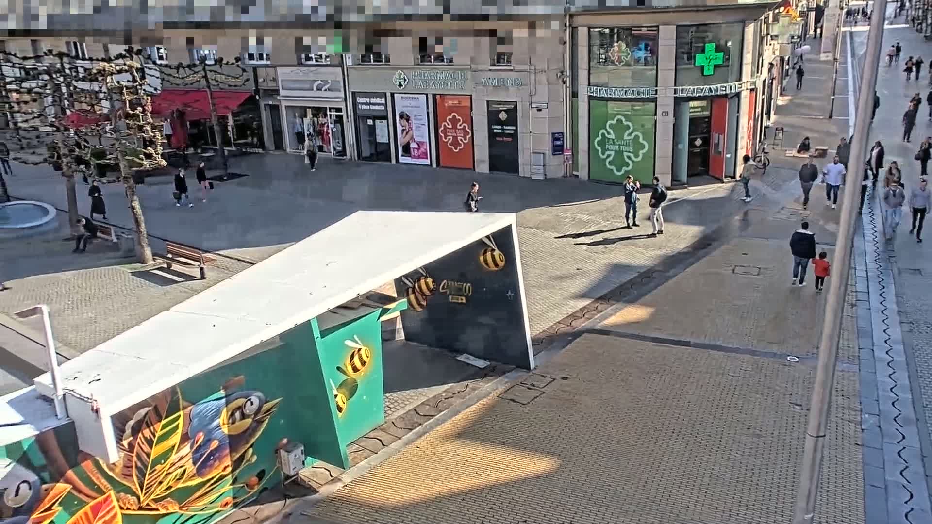 A sunny city square with pedestrians and shops, featuring a modern art installation depicting bees.