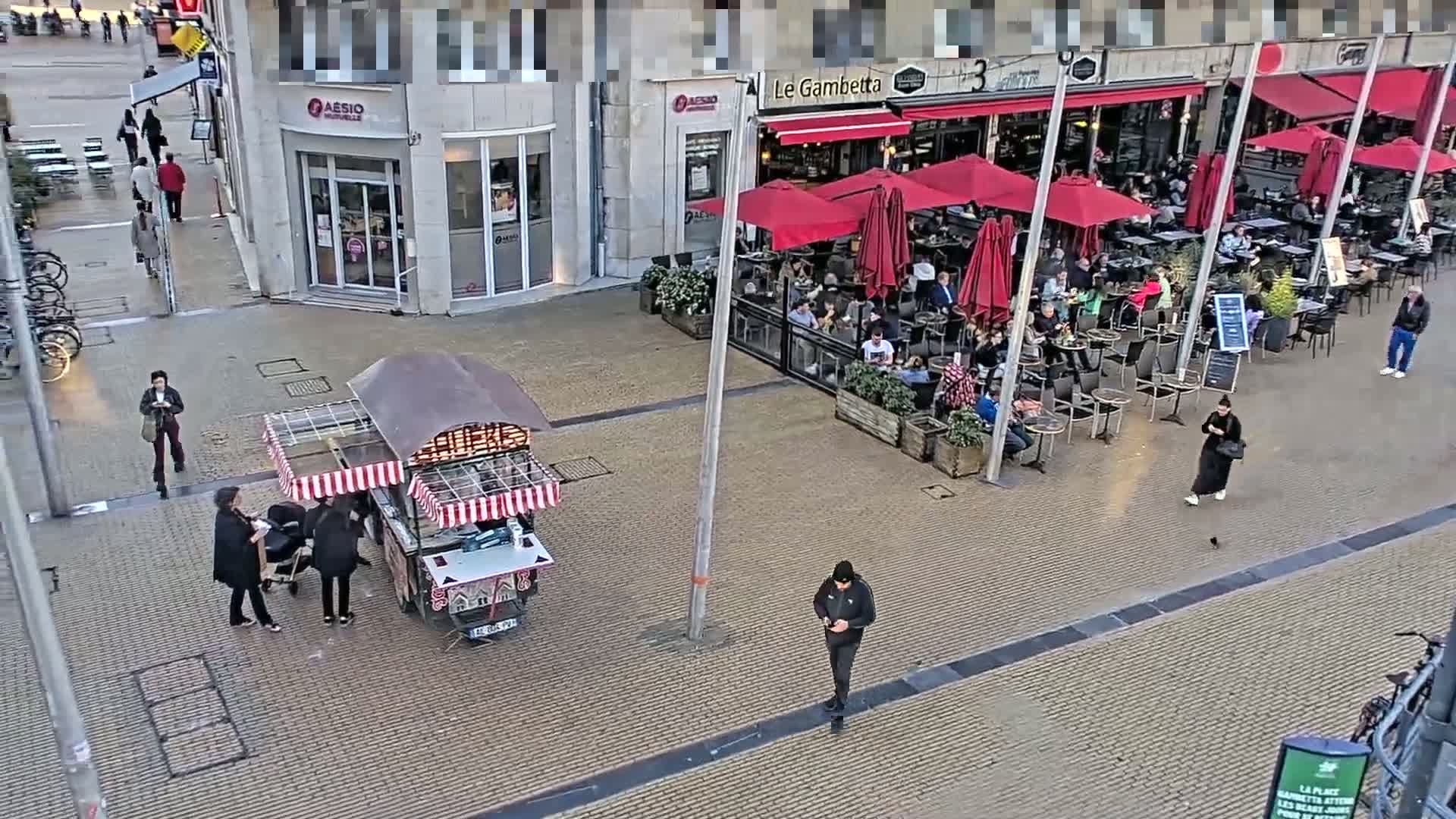 A paved outdoor area shows a food cart, several pedestrians, and an outdoor cafe with red umbrellas under an overcast sky.