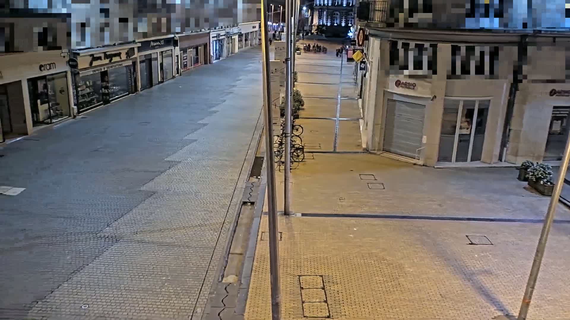 A mostly empty, paved street at night is lined with shops, some with their shutters down, and a few bicycles are parked near a light post.