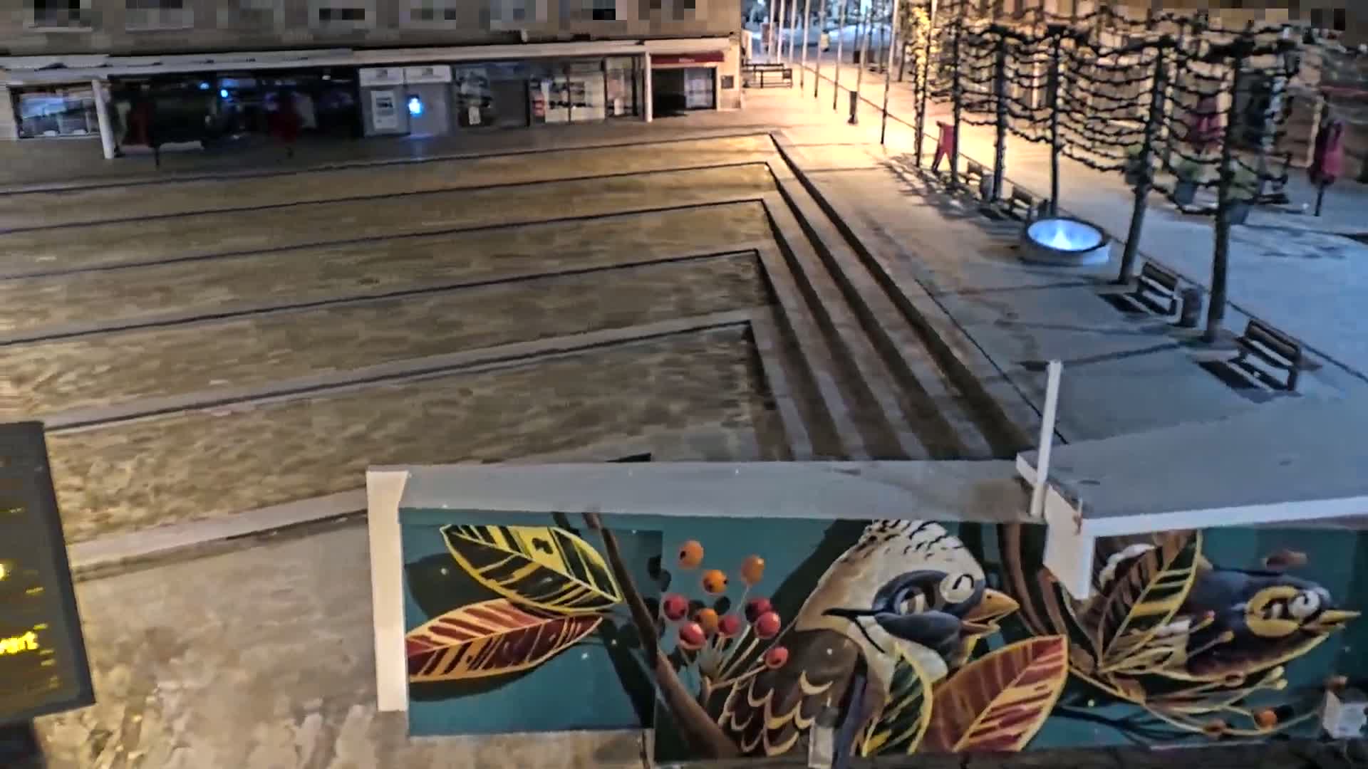 A nighttime view of a plaza with a water feature, benches, and a mural depicting birds and foliage.
