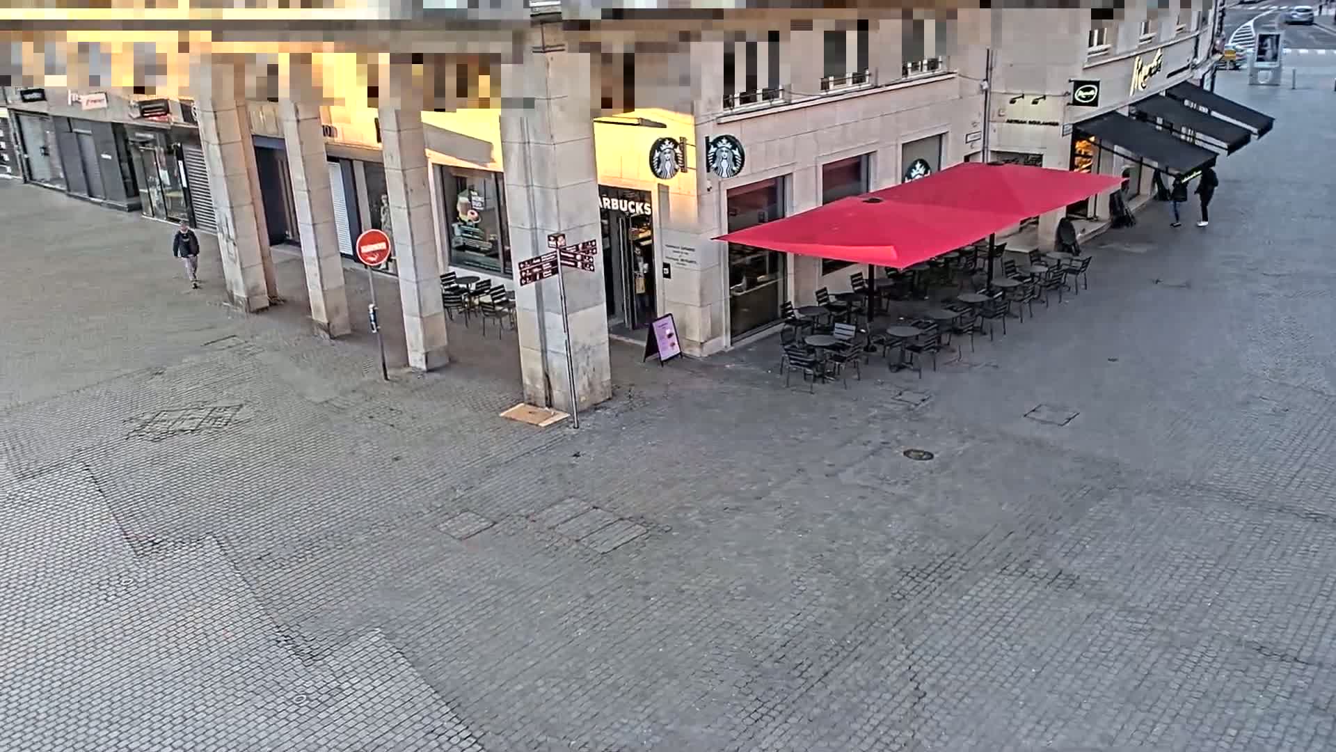 A paved plaza features a Starbucks with a red patio umbrella and several tables and chairs, alongside a pedestrian walking away from the camera, all under a bright but somewhat overcast sky.