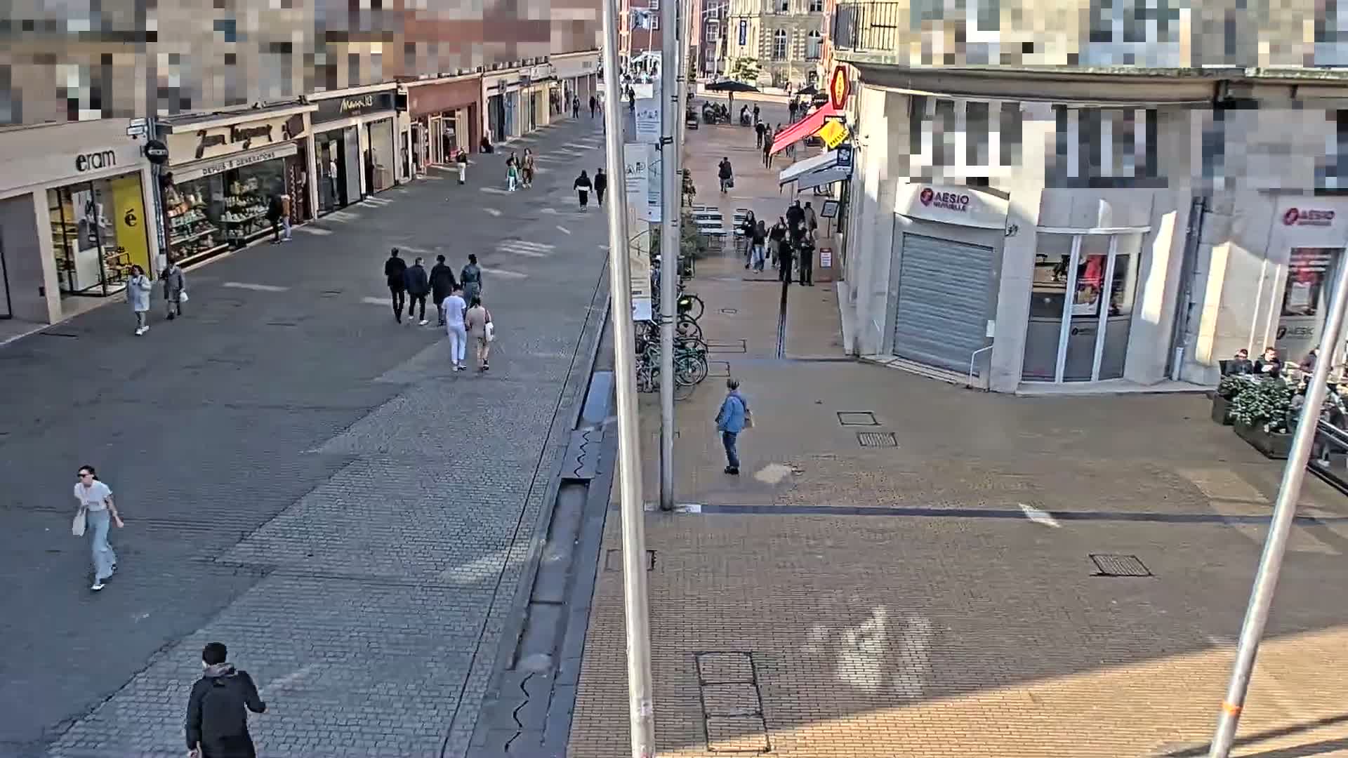 A mostly sunny pedestrian shopping street with several people walking past shops and cafes.