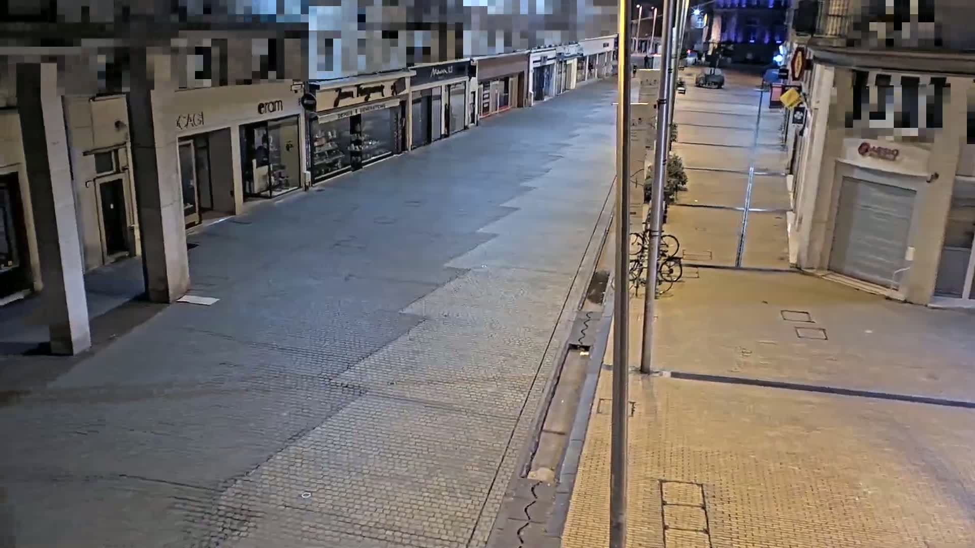 A mostly empty street at night, lined with storefronts, is illuminated by streetlights.