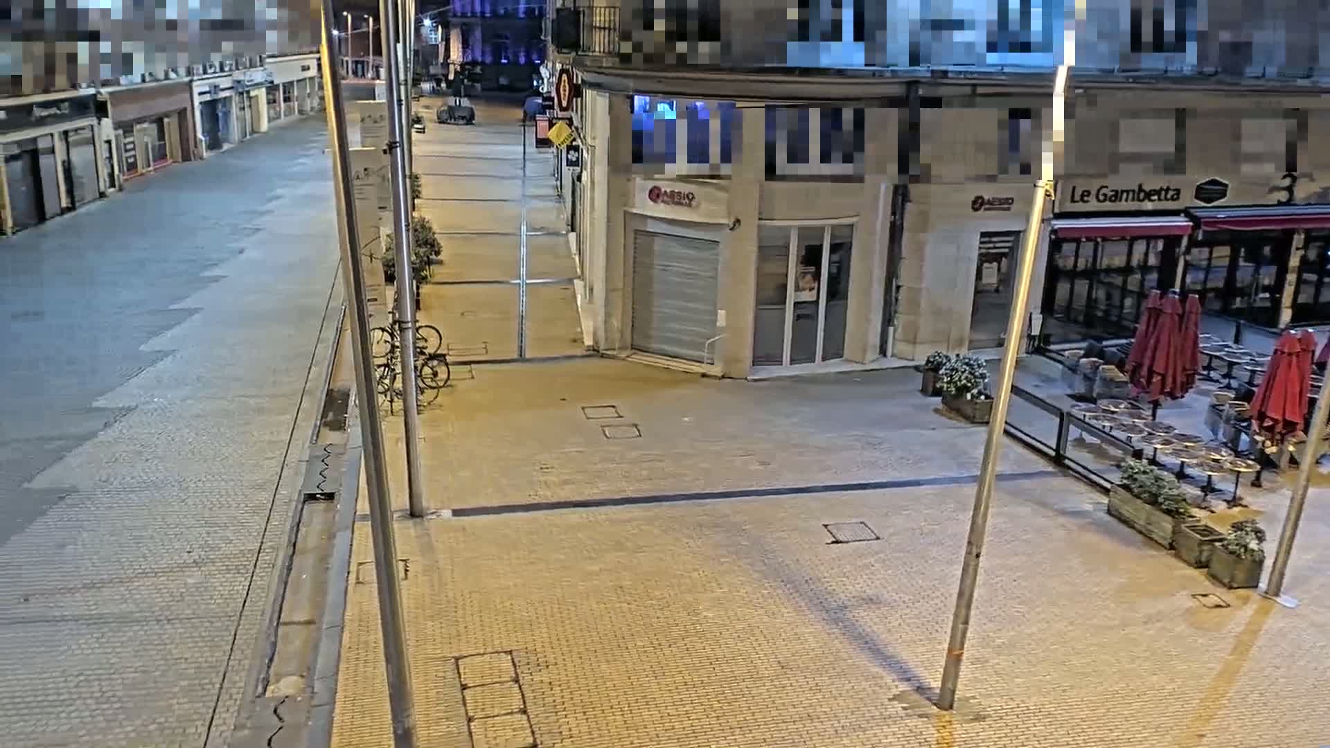 A mostly empty, paved street at night, with two bicycles parked near a building and closed outdoor seating area.