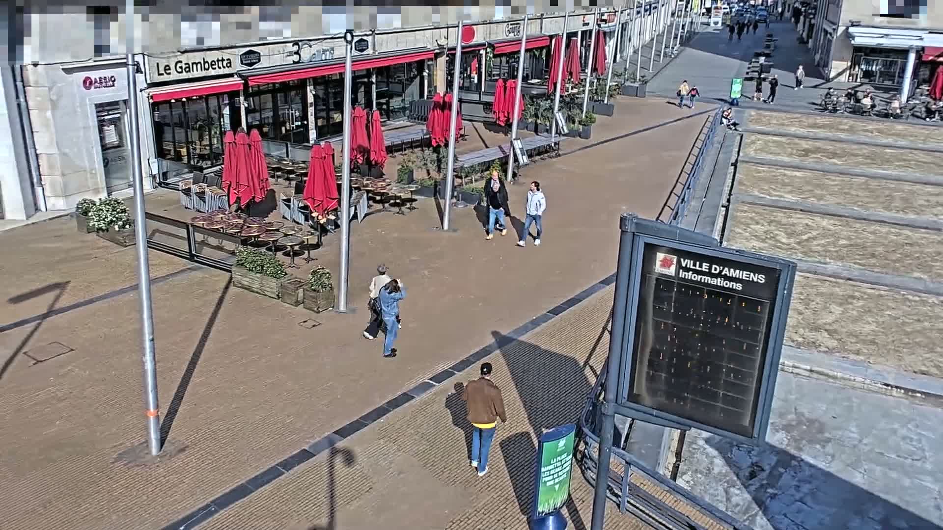 A sunny outdoor plaza with several people walking past red patio umbrellas and tables outside of shops.