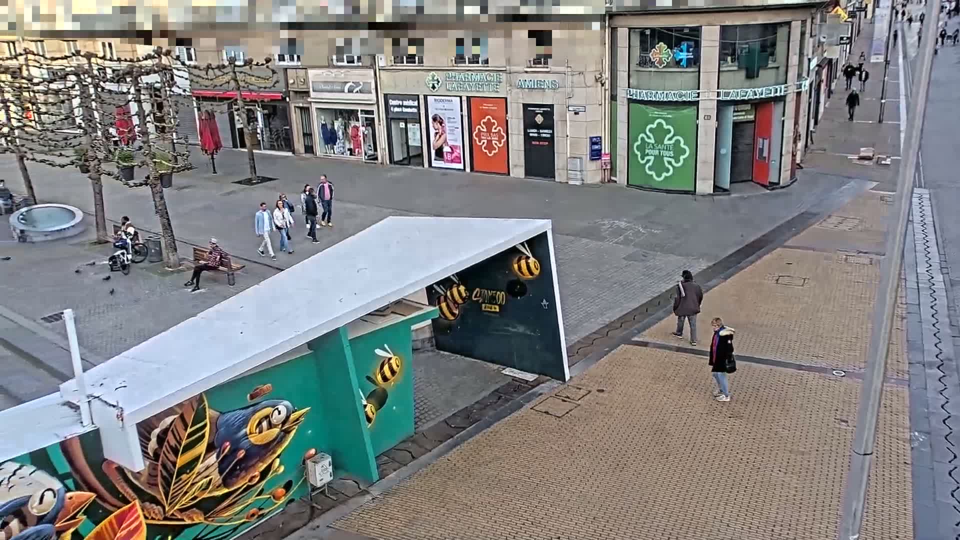 A mostly empty paved city square with several pedestrians, a bicycle, and a mural depicting birds and bees, under a partly cloudy sky.