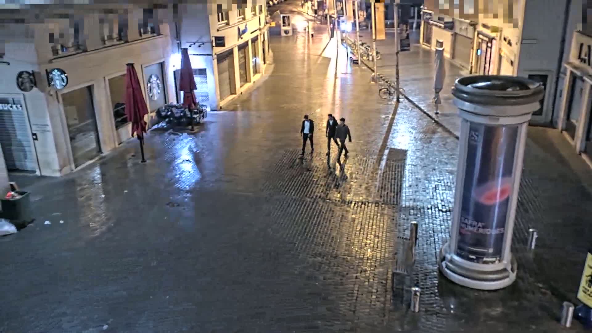 Three men walk down a rain-slicked cobblestone street at night past buildings with closed storefronts.