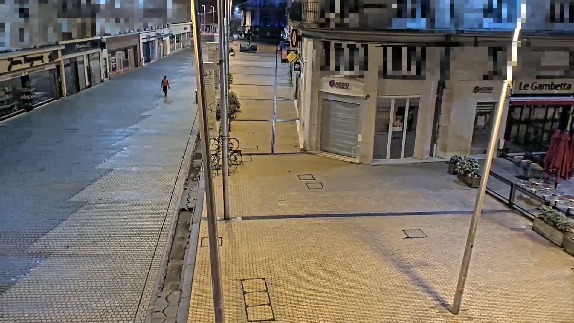 A person walks down a mostly empty, paved street at night, past shops and a restaurant with outdoor seating, under well-lit streetlights.