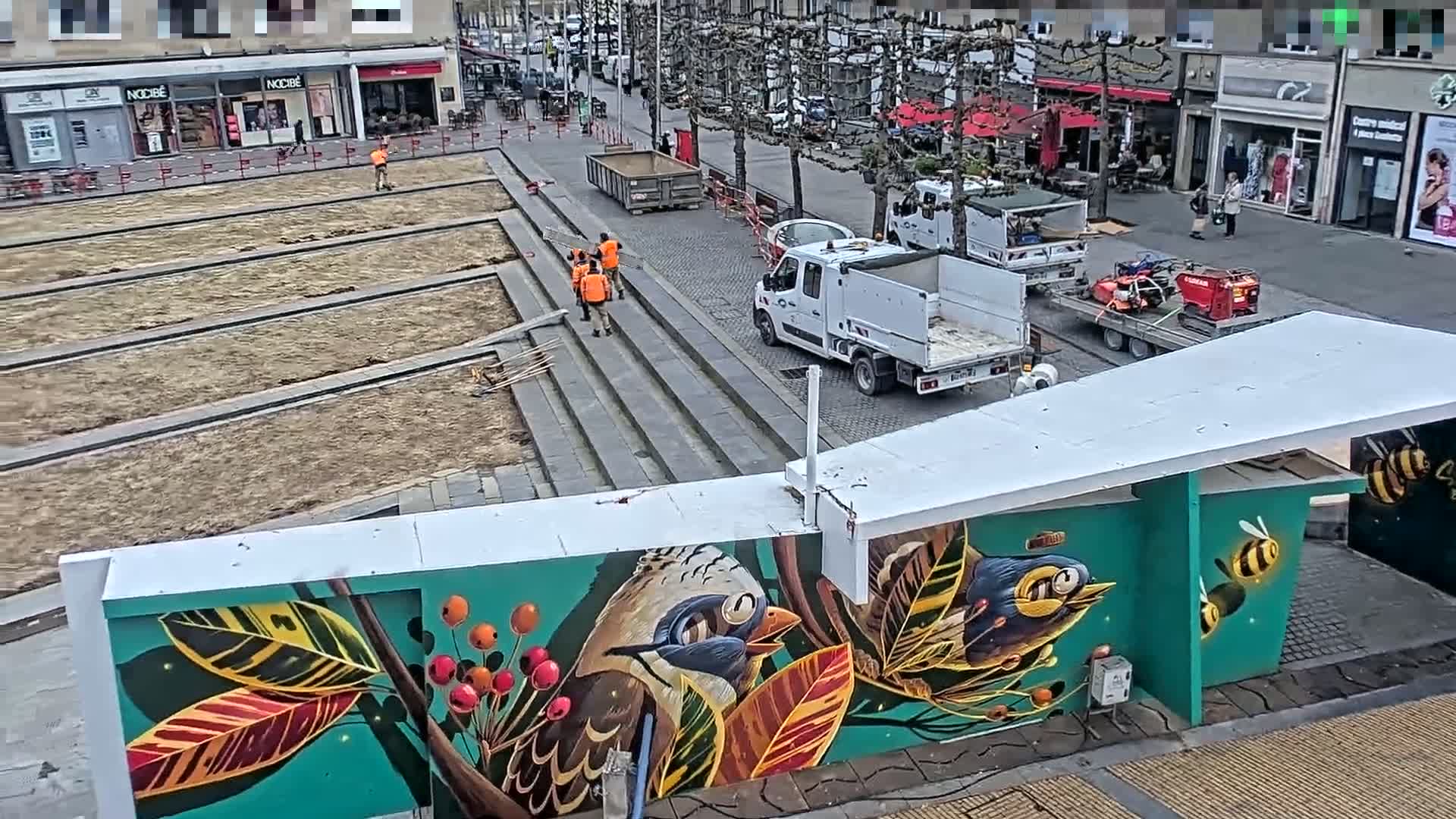 Construction workers are working on a public space featuring a vibrant mural of birds and foliage, under a partly cloudy sky.