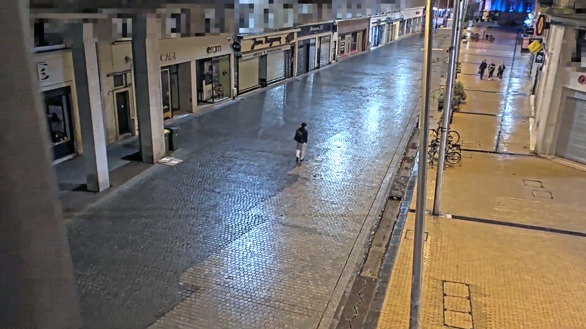 A lone person walks down a wet, cobblestone street at night, past closed shops and parked bicycles.