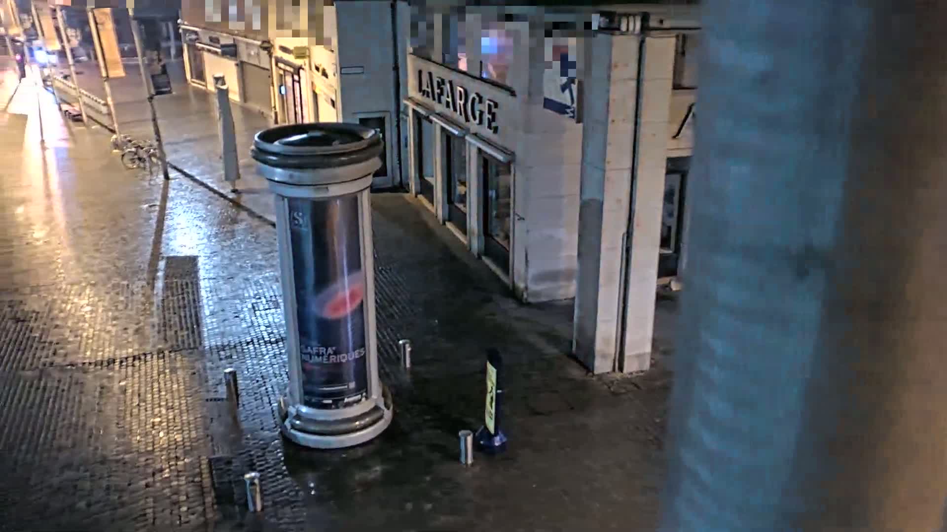 A wet cobblestone street at night is seen from an elevated vantage point, showing a cylindrical advertisement stand and a building in the background.