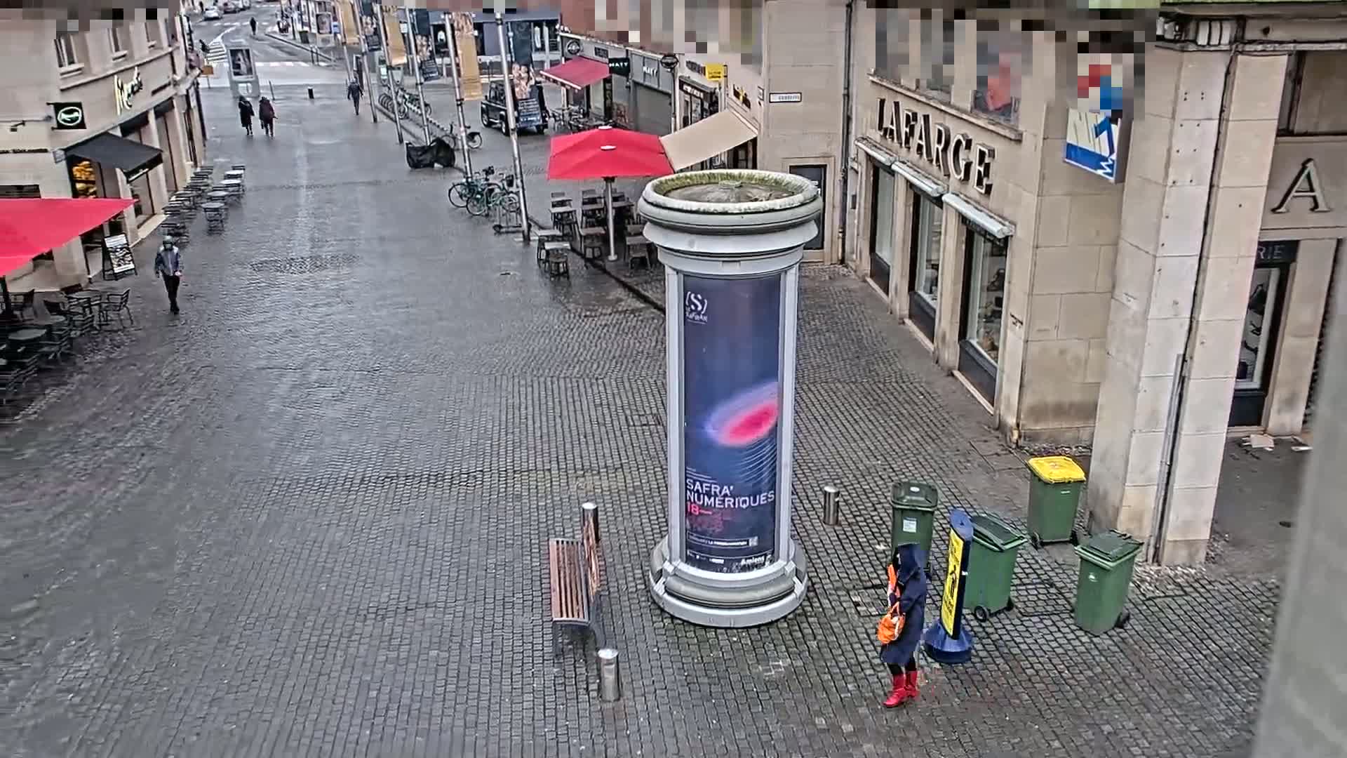 A wet cobblestone street scene shows a tall column with an advertisement, a woman in red boots, several trash cans, and outdoor seating areas under red umbrellas.