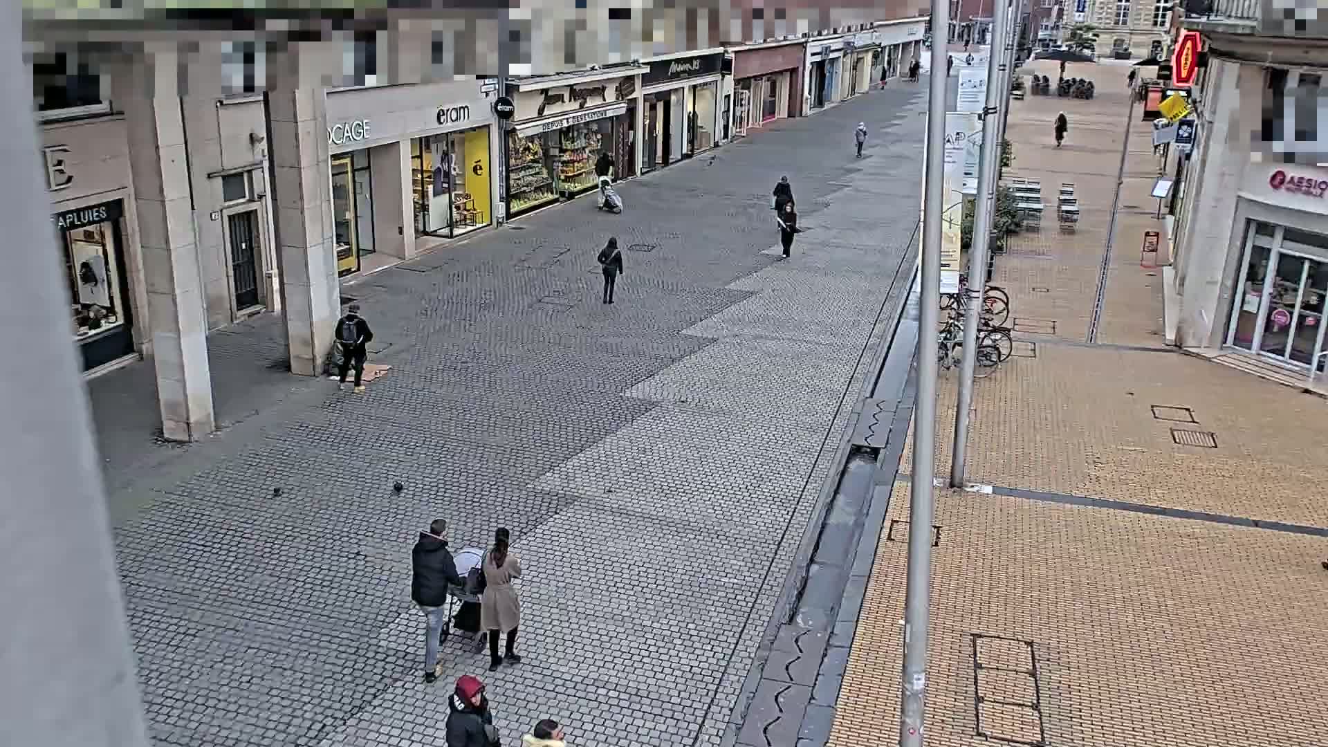 A cobblestone street lined with shops, mostly empty except for several pedestrians and a small group of people near a stroller.