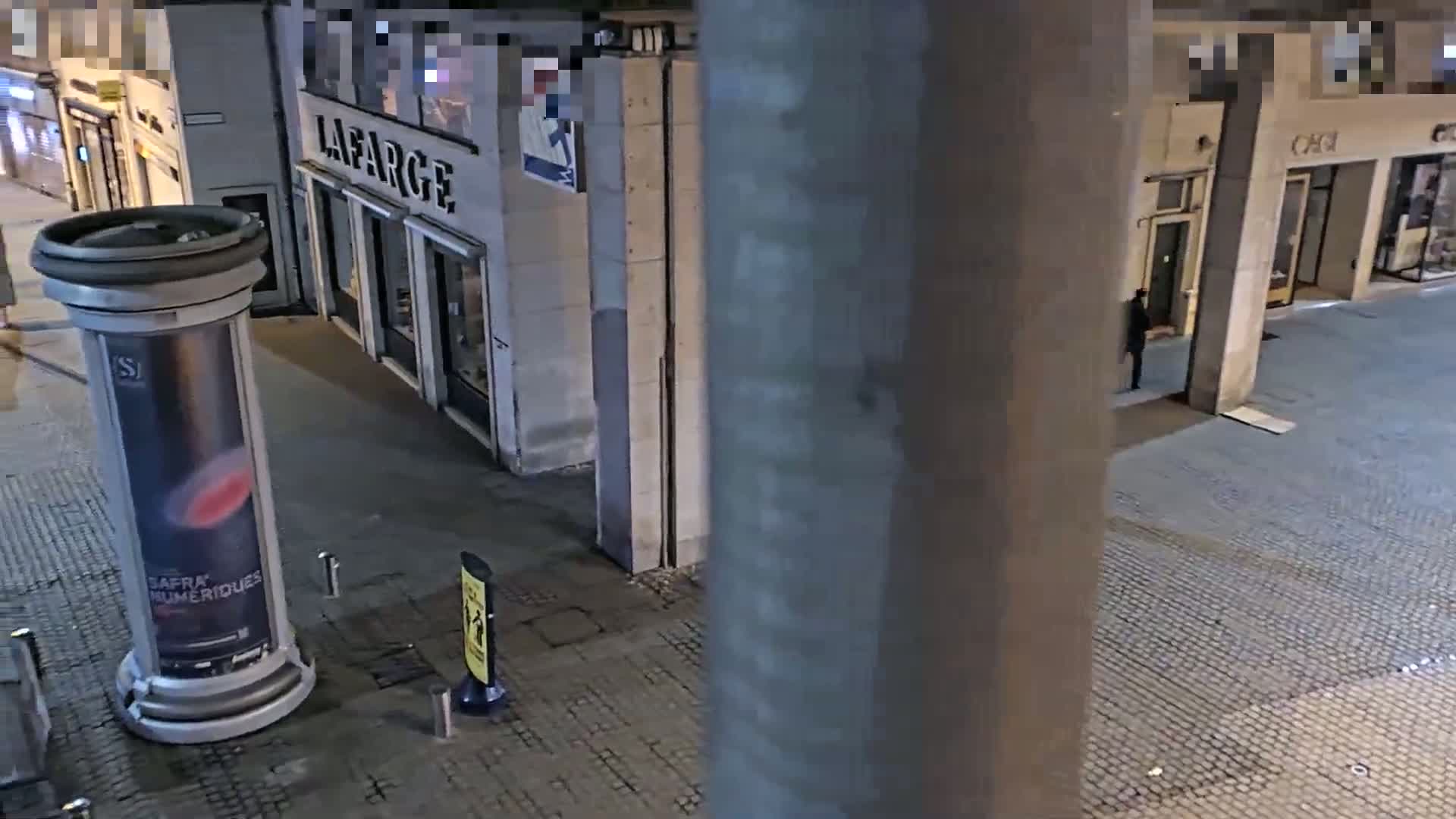 A nighttime, high-angle view shows a cobblestone street with several storefronts, a large advertisement pillar, and a lone pedestrian.