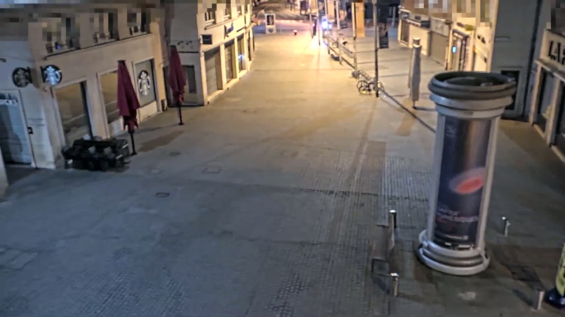 A mostly empty street at night is seen from above, with closed shops and a few streetlights illuminating the pavement.