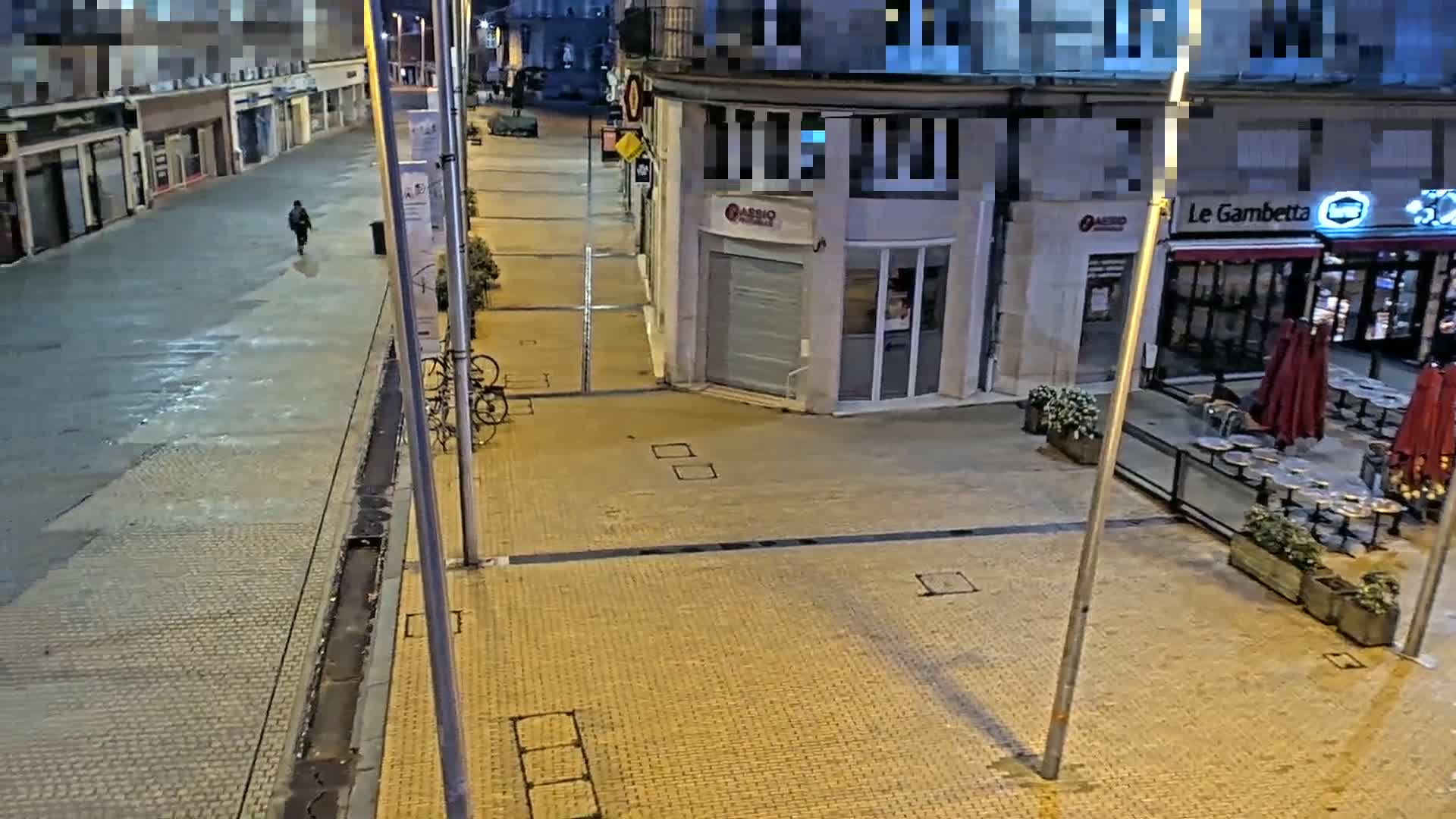 A mostly empty, paved city street at night, with a lone pedestrian walking away from the camera, shows a few parked bicycles, and a restaurant with its outdoor seating area closed.