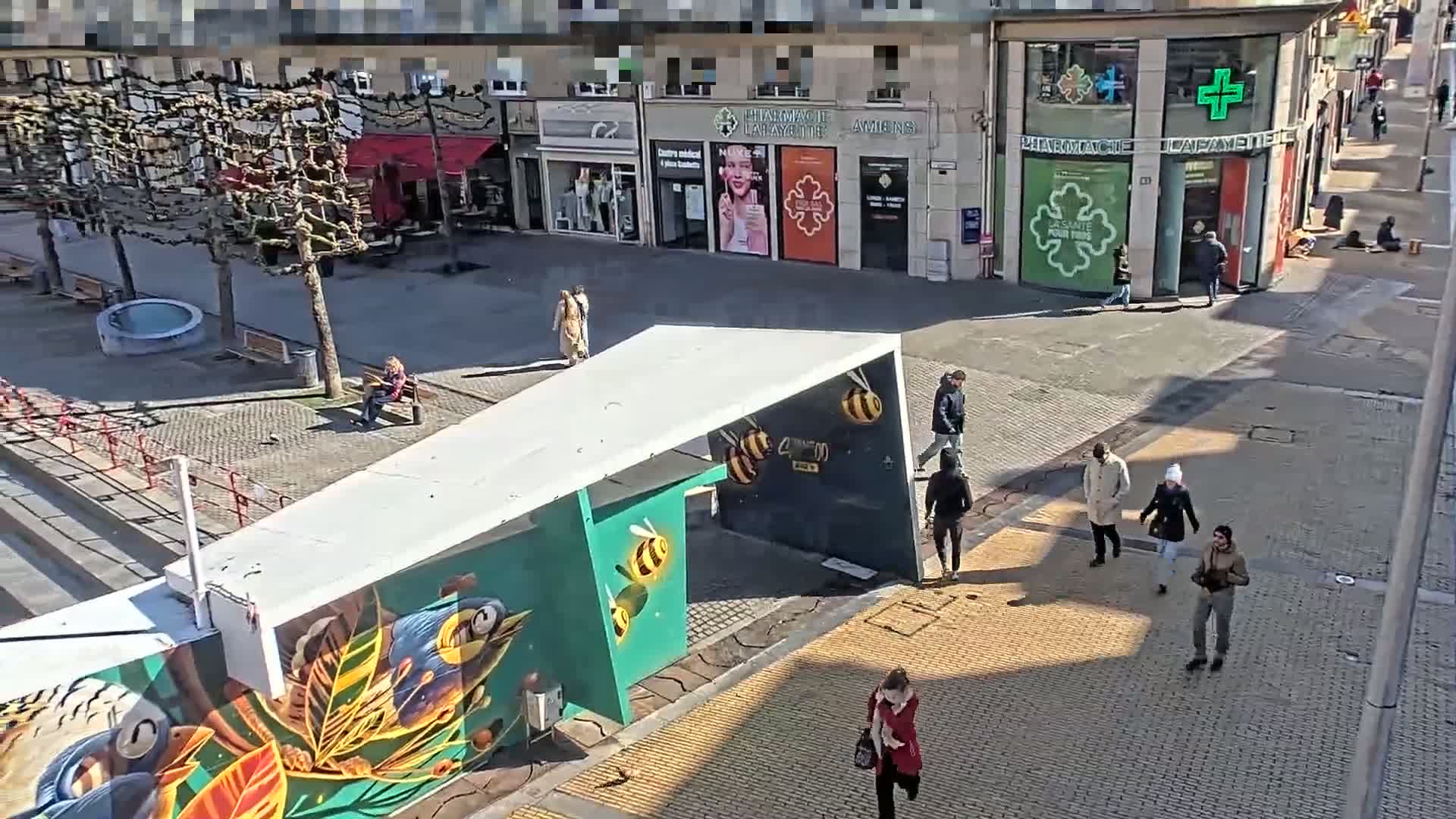 A sunny city square with several pedestrians walking past a modern structure decorated with a mural of birds and bees.