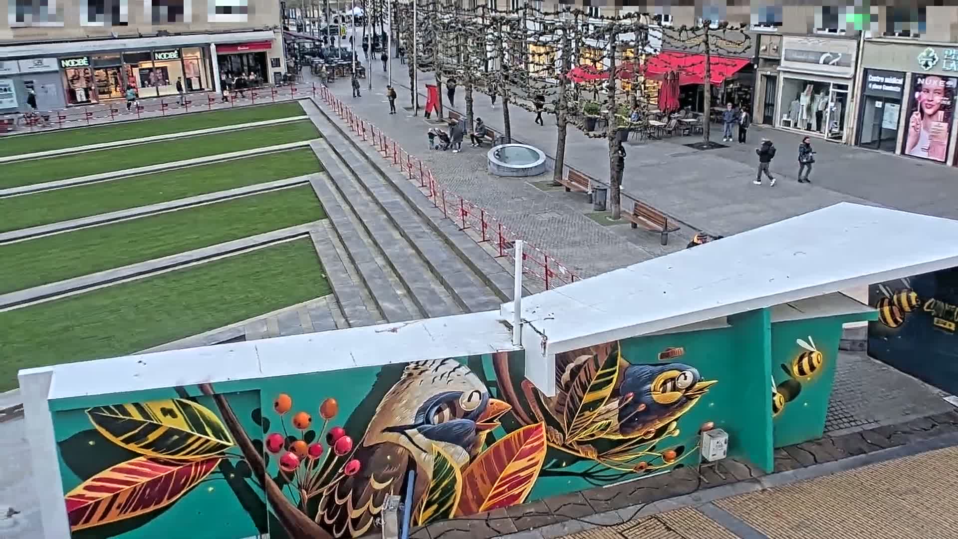 A vibrant mural depicting birds and bees adorns a wall in a city square, where several pedestrians stroll under a mostly clear sky.