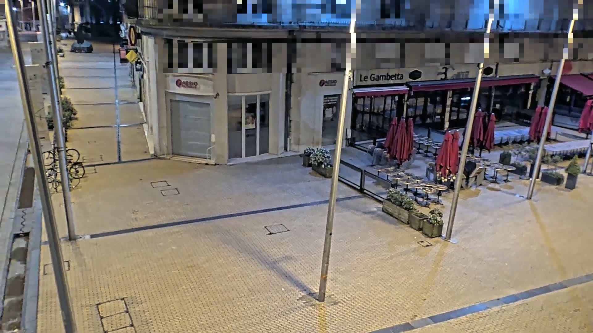At night, a paved area outside a restaurant with closed red umbrellas and empty tables is shown, with a few bicycles parked nearby.