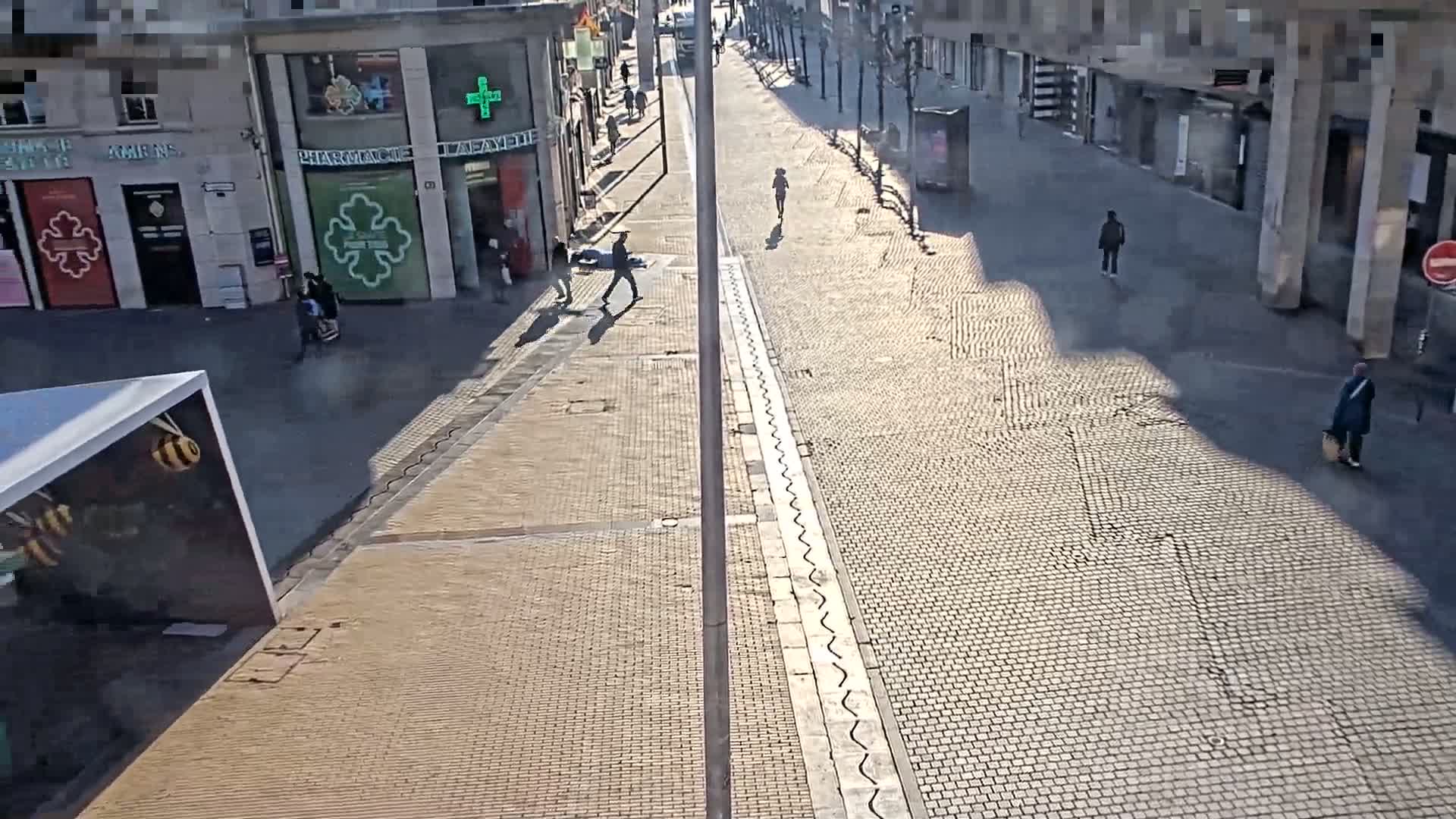 A sunny day reveals a mostly empty cobblestone street with several pedestrians and buildings lining both sides.