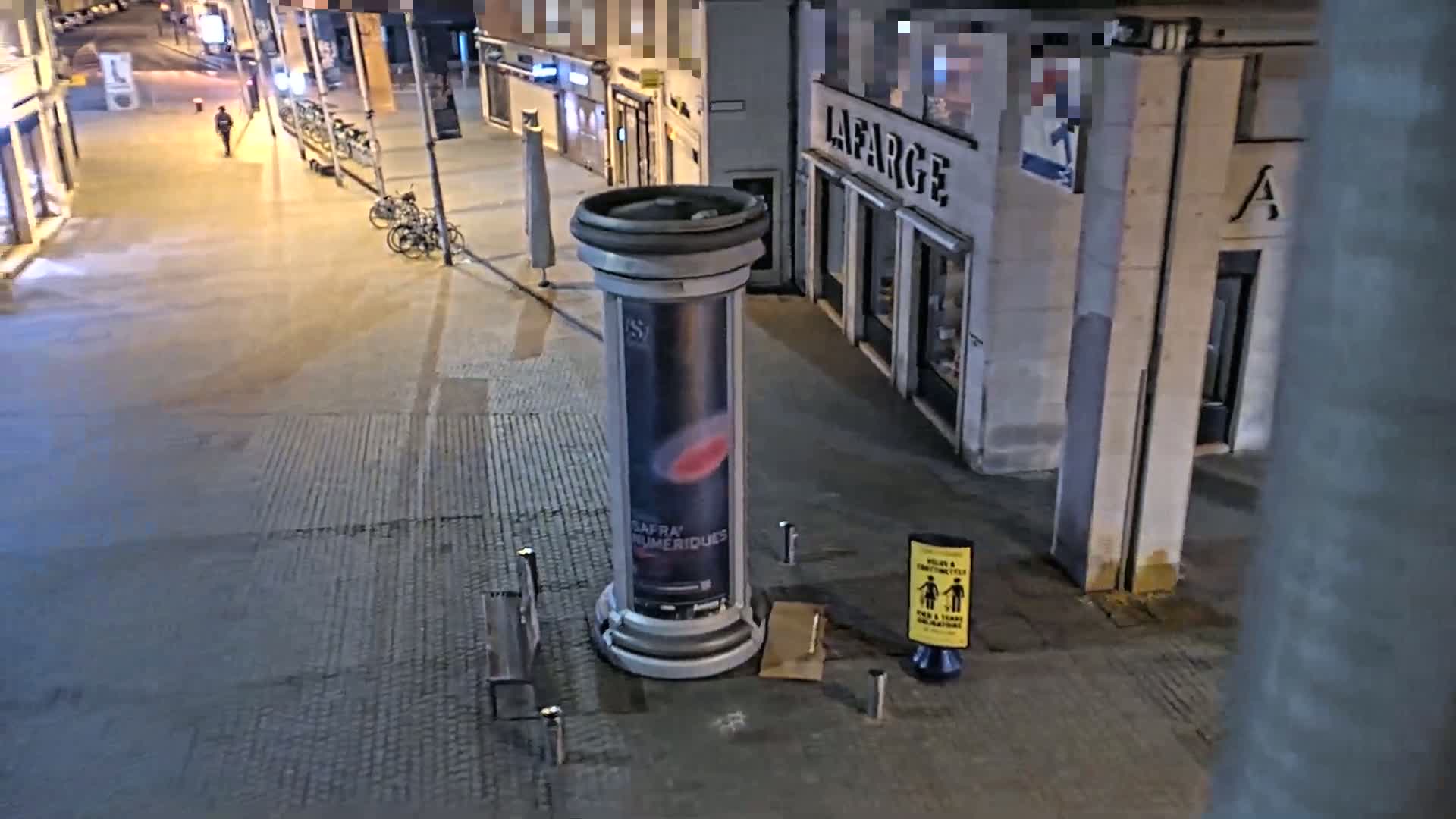 A nighttime, city street scene shows a large advertising column with a poster, two benches, a couple of bicycles, and a lone pedestrian walking in the distance.