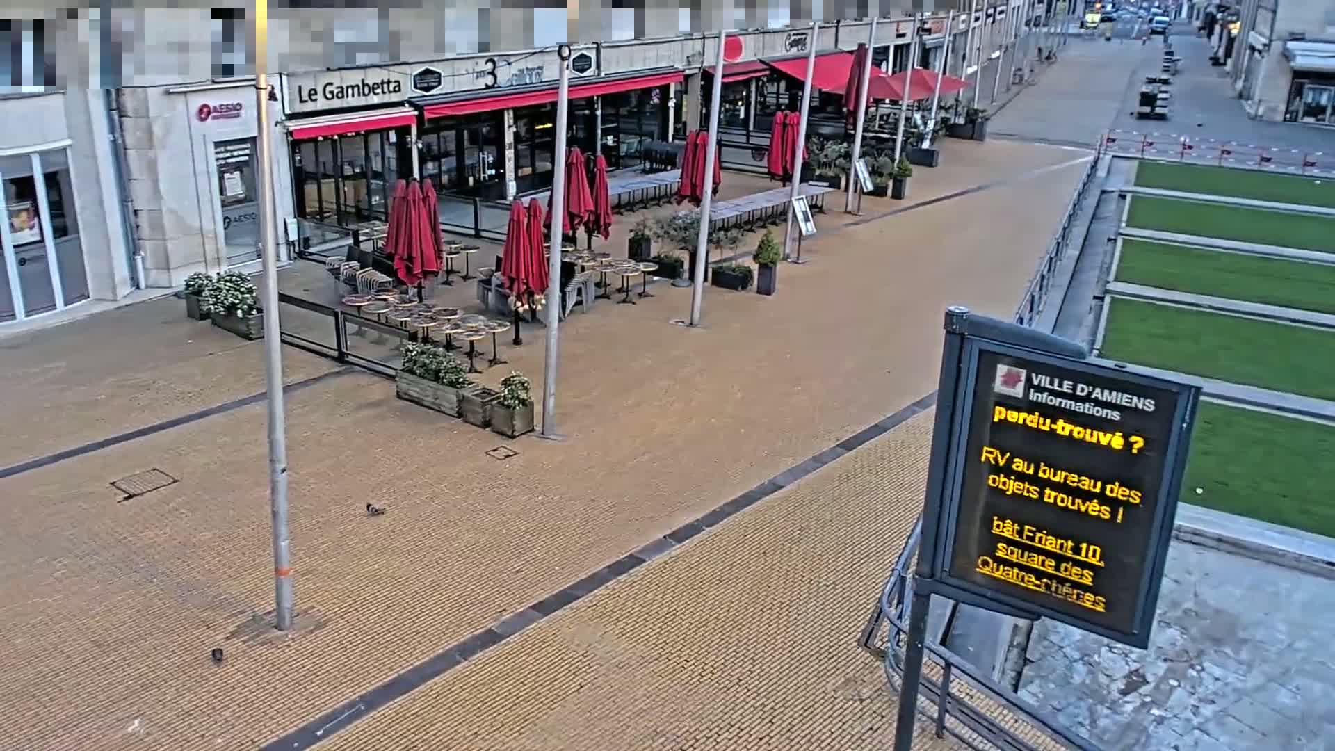 An outdoor cafe with red umbrellas and tables sits empty in a paved plaza next to a grassy area.