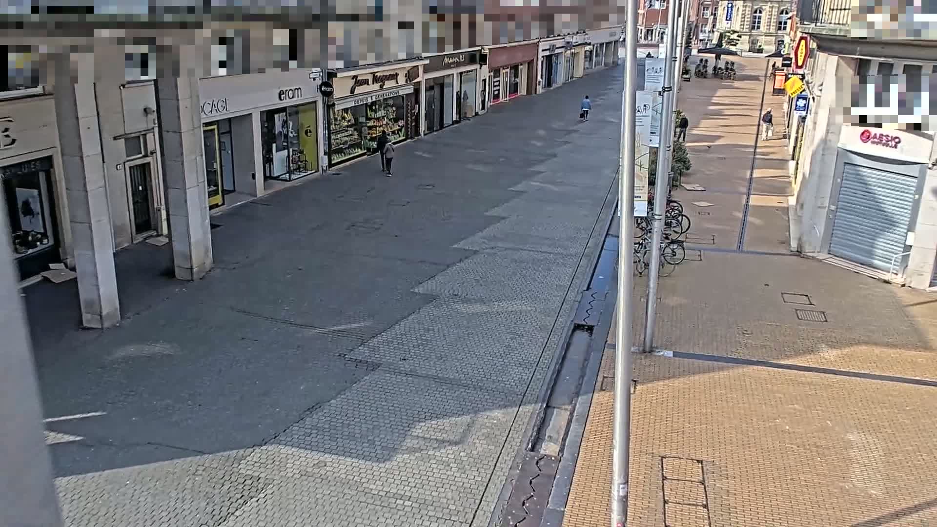 A mostly empty, sunlit pedestrian shopping street with several closed shops and a few people walking, lined with buildings and some bicycles parked at the side.