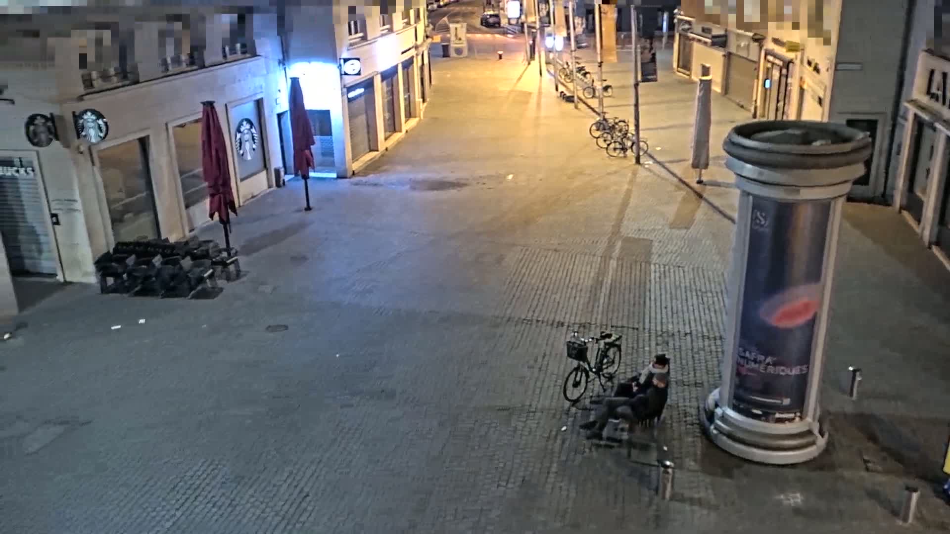 A person sits on a bench next to a bicycle in a mostly empty city street at night.