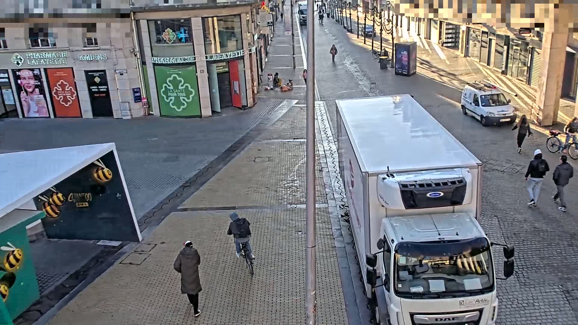 A sunny city street scene shows several pedestrians and cyclists walking and biking past a large white refrigerated truck parked at the curb, near buildings with storefronts.