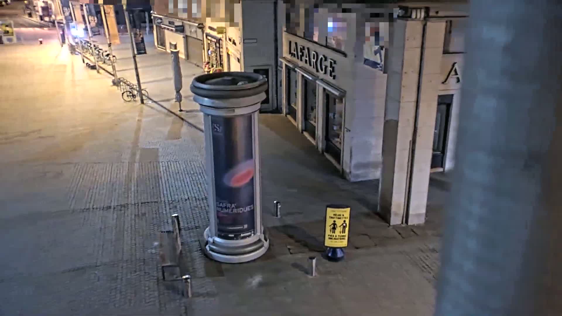 A nighttime street scene shows a cylindrical advertisement stand next to a building, with a few parked bicycles visible in the background.