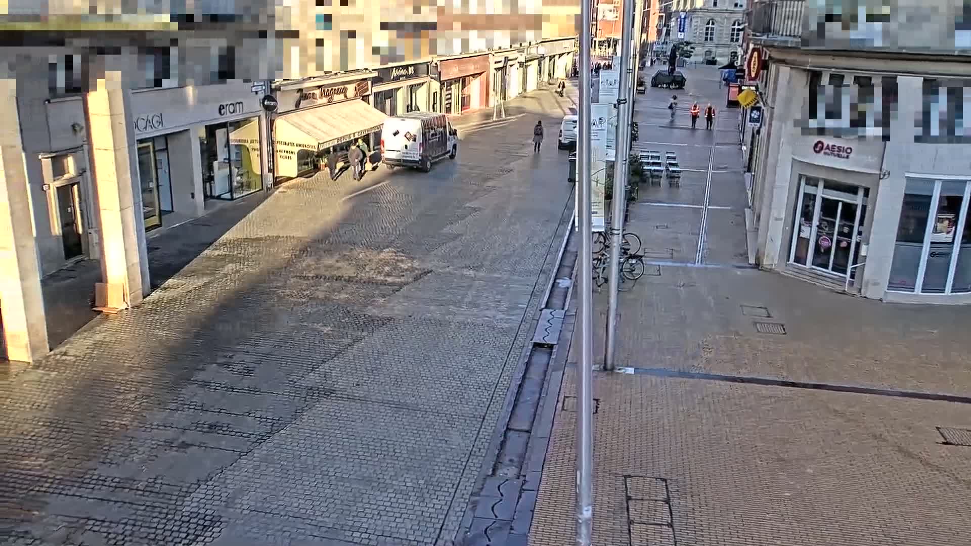 A cobblestone street scene shows several pedestrians and a delivery van in front of shops on a sunny day.