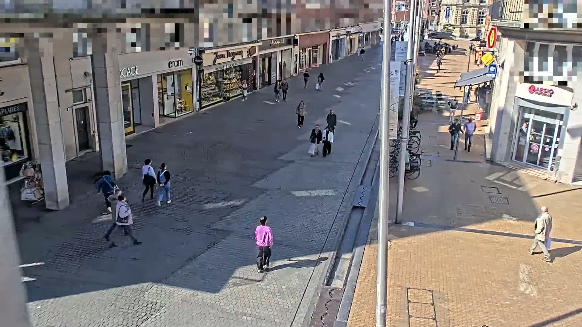 A sunny day on a cobblestone pedestrian street shows several people walking past shops and buildings.