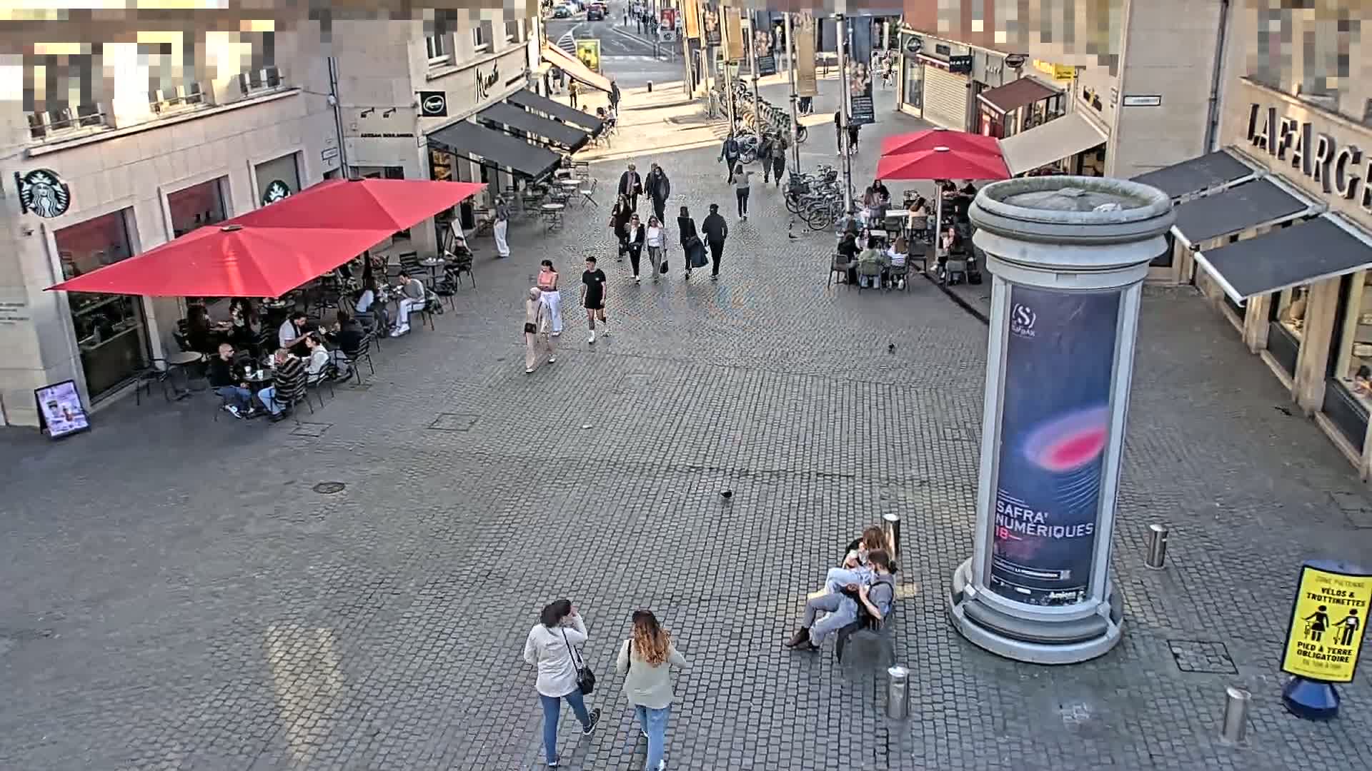 A cobblestone plaza with several people walking and sitting at outdoor cafes under red umbrellas on a sunny day.