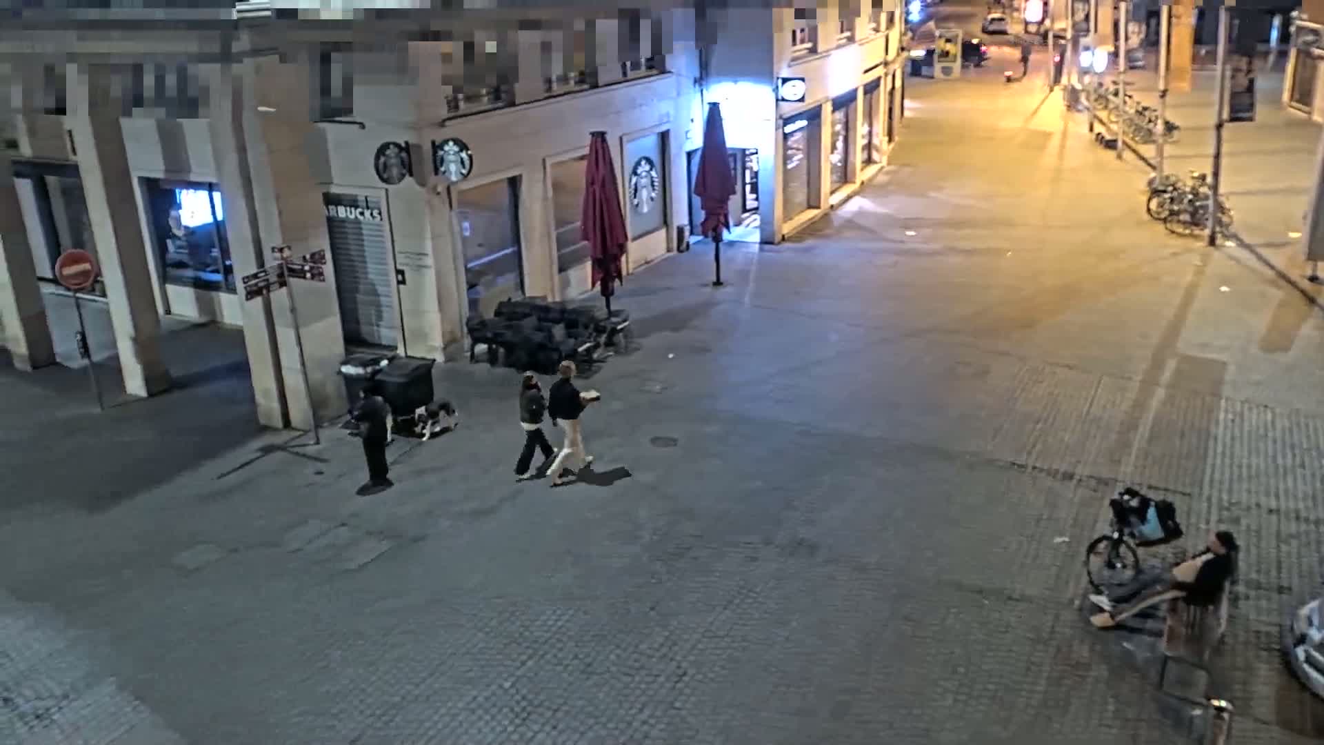 At night, several people walk near a Starbucks and a person sits on a bench next to a bicycle in a paved city area.
