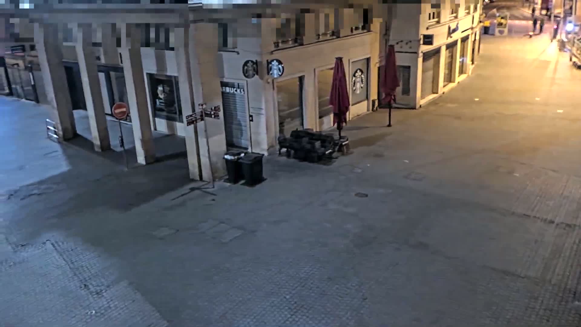 A nighttime street scene shows a mostly empty paved area in front of a building with closed storefronts, including a Starbucks, and a couple of closed umbrellas.