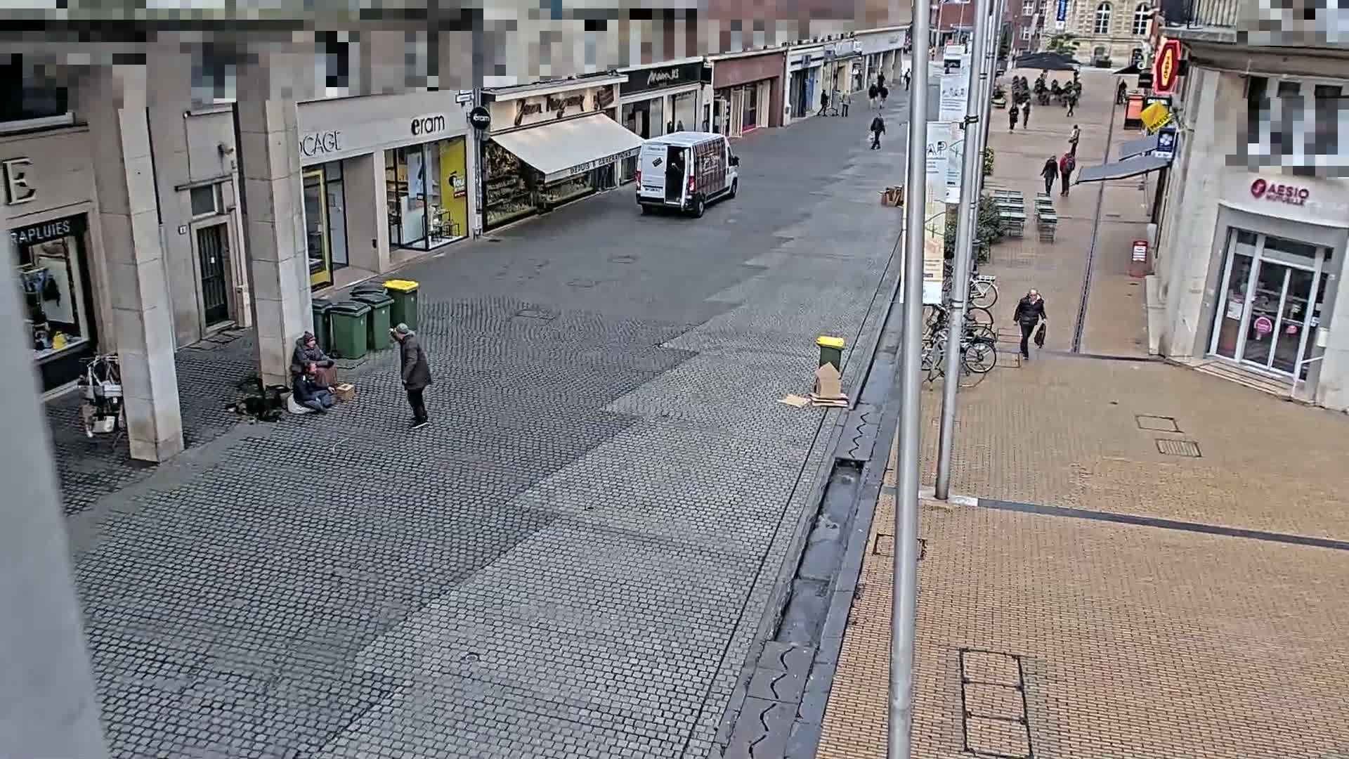 A cobblestone street with shops and buildings on either side, featuring a few pedestrians and two people sitting on the ground near some trash cans.