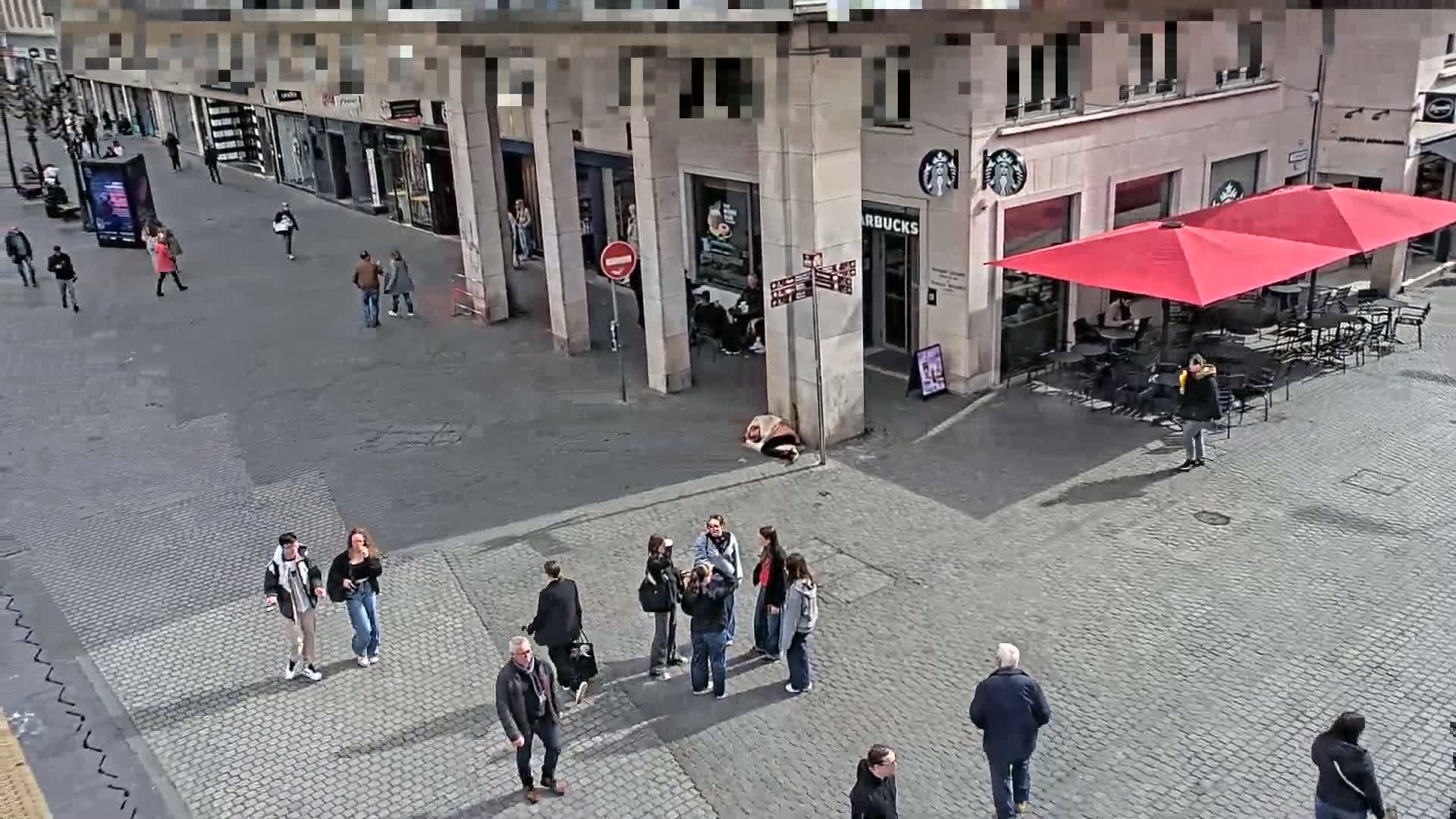 A sunny city street scene shows pedestrians walking past shops and a Starbucks with red umbrellas, where several people are seated at outdoor tables; a person is lying on the ground near the Starbucks.