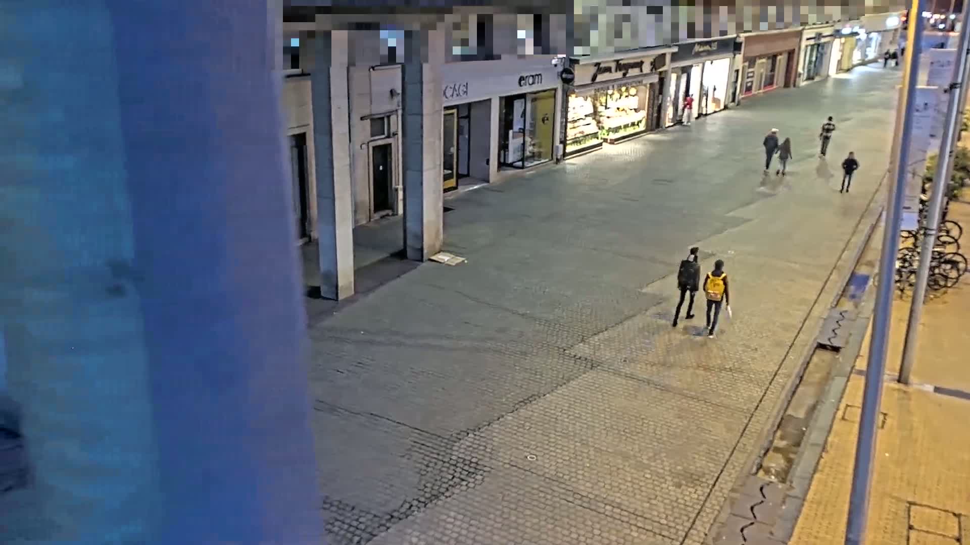 A nighttime street scene shows several pedestrians walking past shops and a row of parked bicycles.