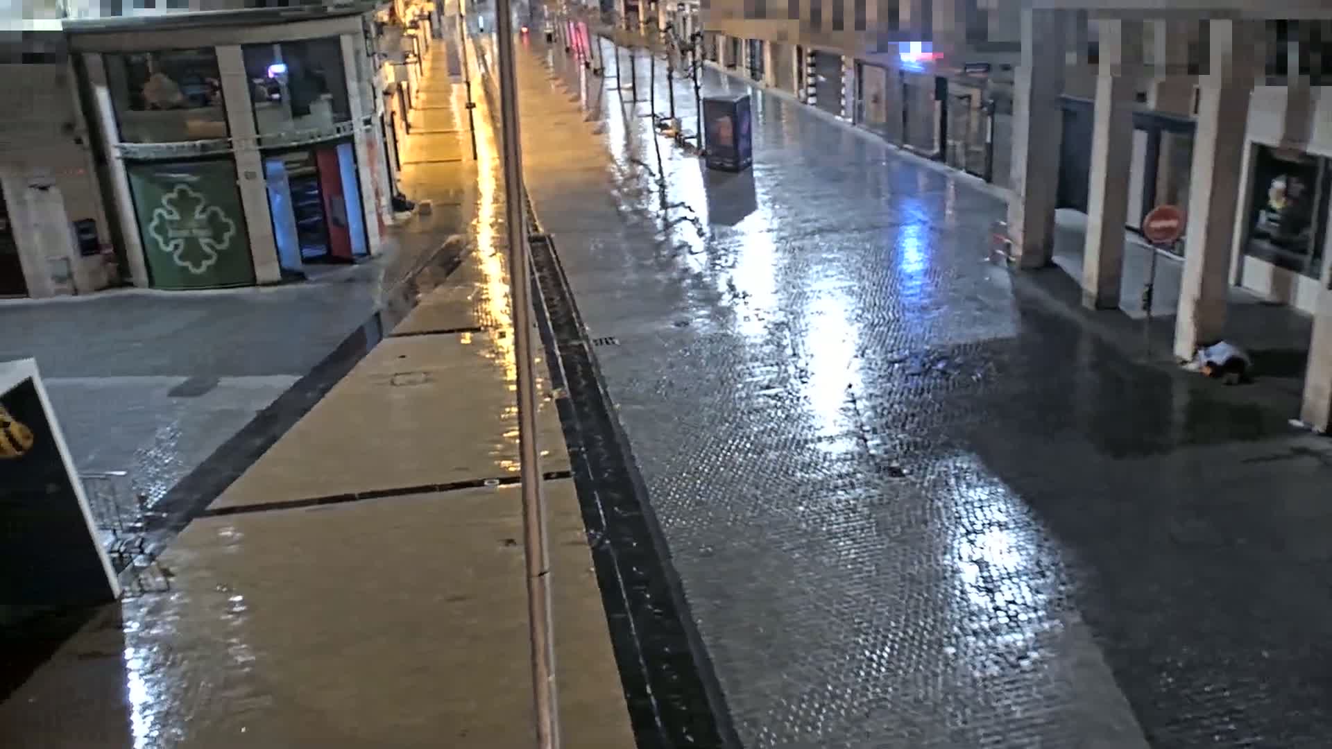 A rain-slicked, empty city street at night is lit by streetlights, reflecting on the wet cobblestones.