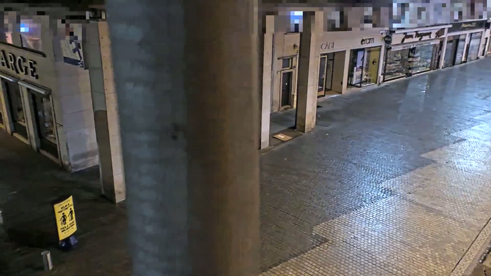 A wet, nighttime street scene shows a row of storefronts under a covered walkway.