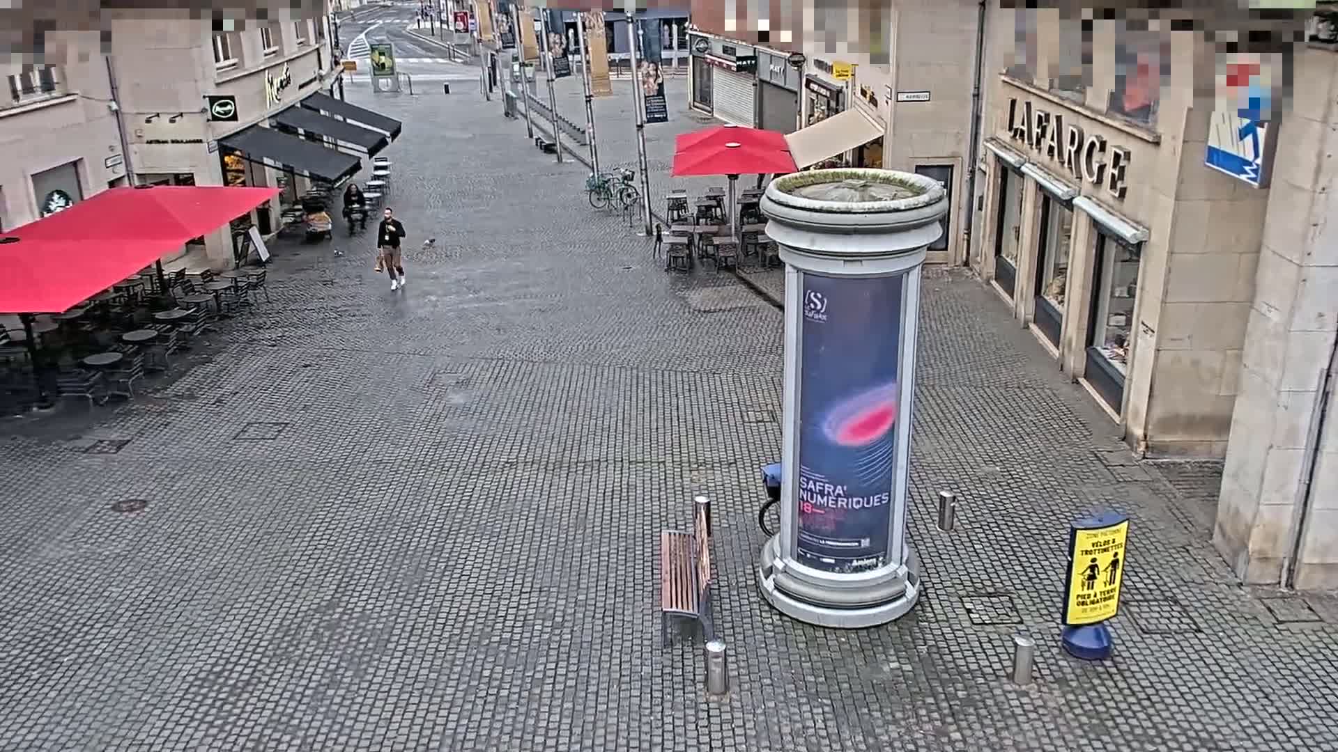 A person walks past an outdoor cafe with red umbrellas on a wet, cobblestone street, near a tall, cylindrical advertisement stand and a bench.