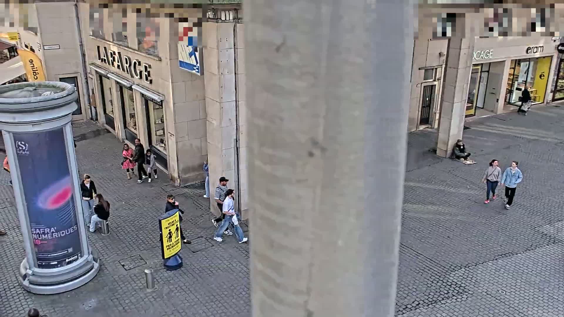 A cobblestone city street scene with several pedestrians, some standing in groups and others walking, under a mostly clear sky.