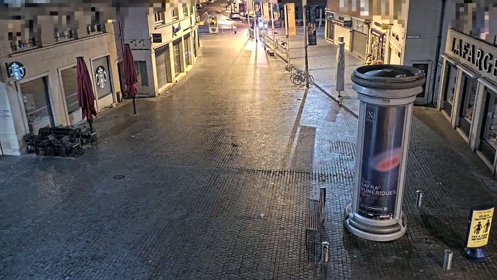 A dark, cobblestone street at night, illuminated by streetlights, shows closed shops and outdoor seating areas with empty chairs.