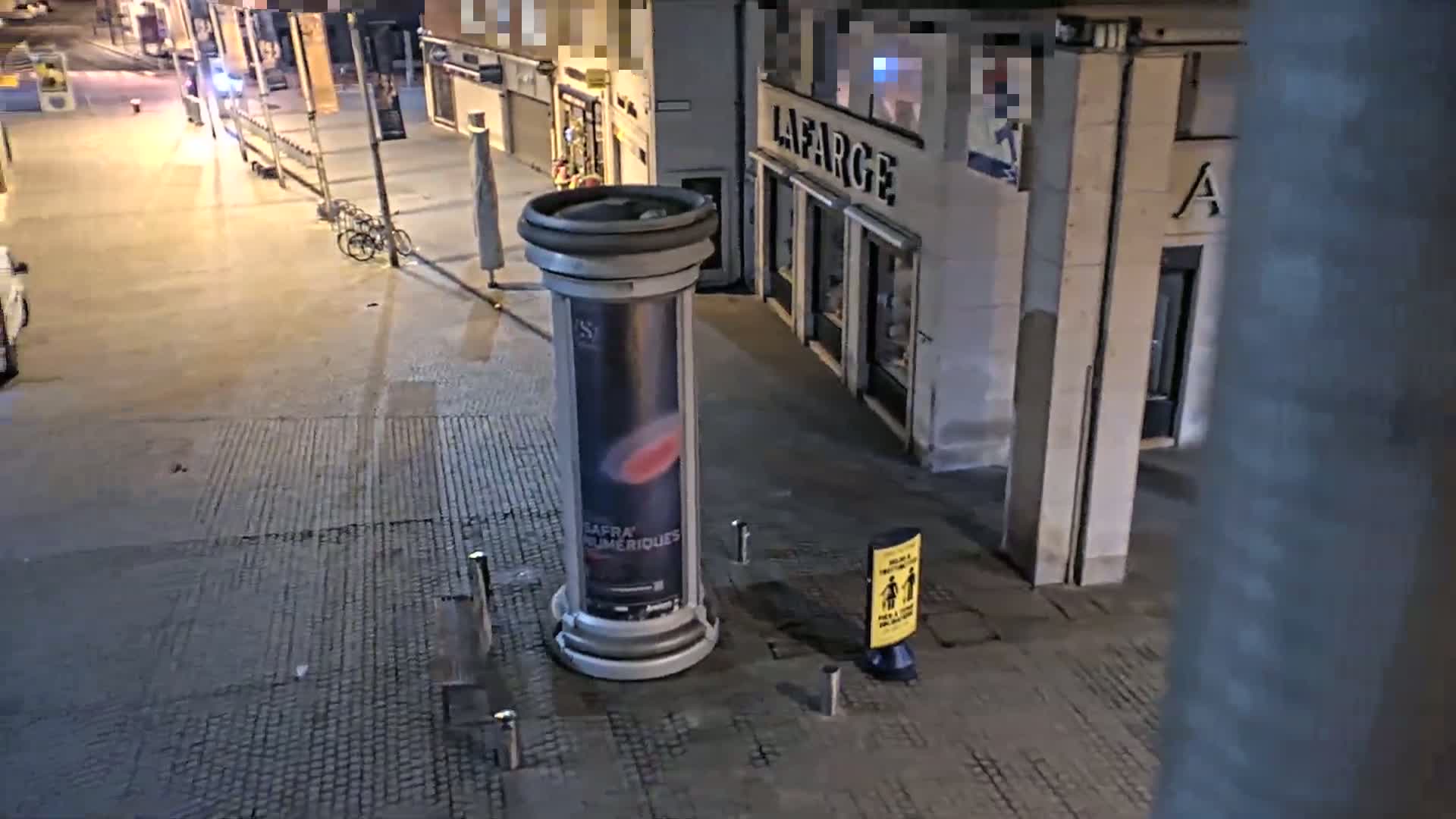 A nighttime street scene shows a paved area with a large advertising column, a bicycle leaning against a railing, and some parked cars, under well-lit conditions.