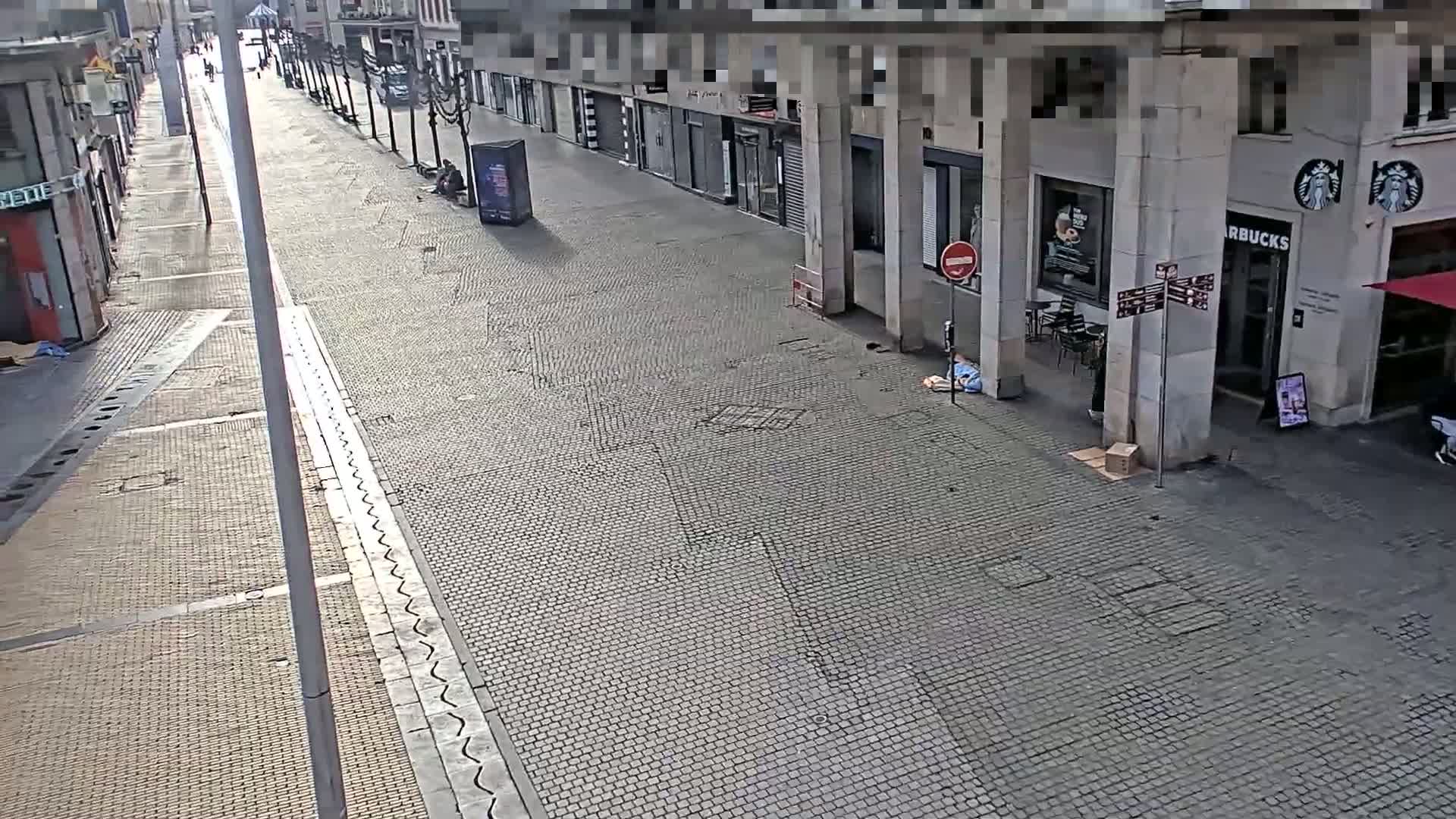 A mostly empty cobblestone street lined with shops on a sunny day, with a single person sitting on the ground near a Starbucks.