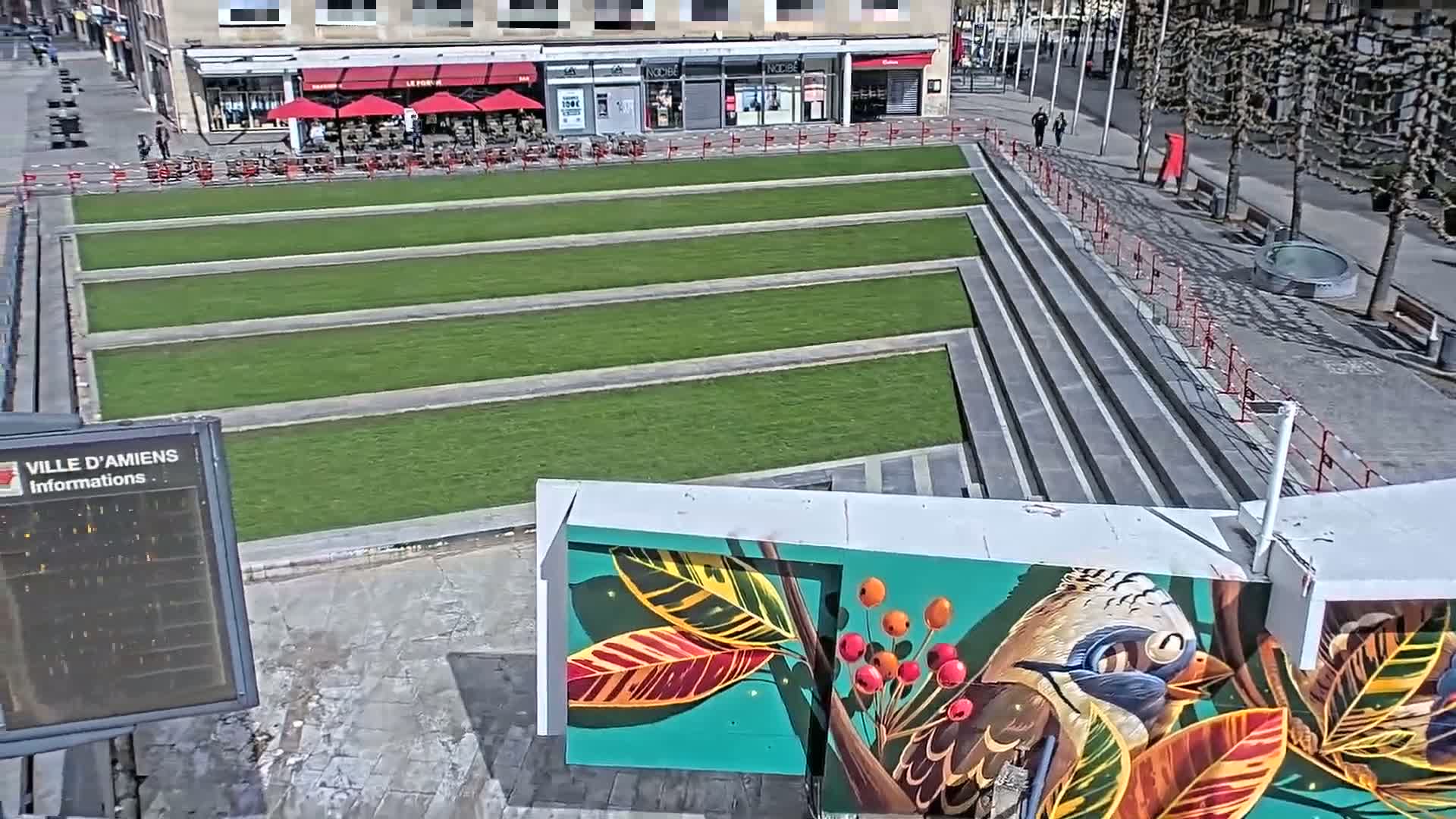 A sunny day reveals a public square with a multi-level grassy area, steps, and a large mural depicting a bird and tropical leaves.