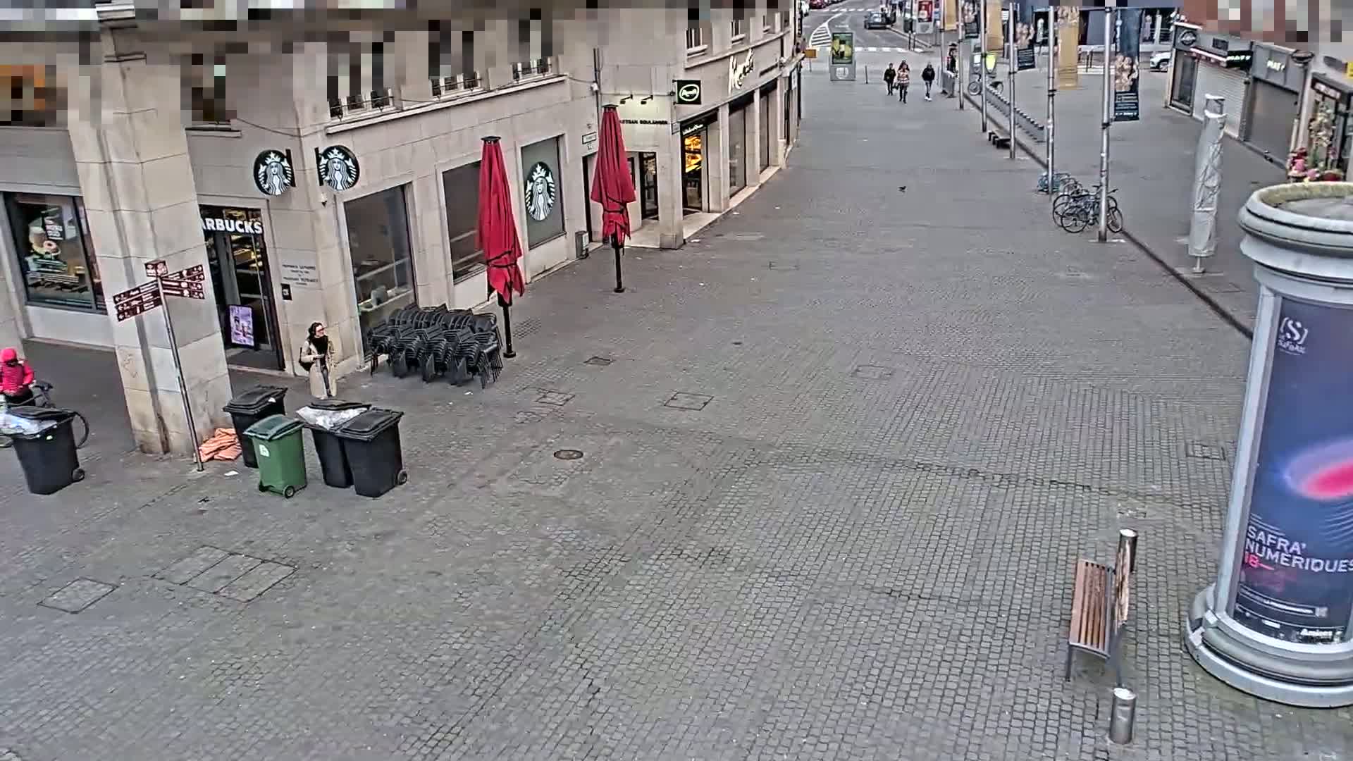 A mostly empty cobblestone street in front of a Starbucks and other shops, with a few people and some stacked chairs outside, under an overcast sky.